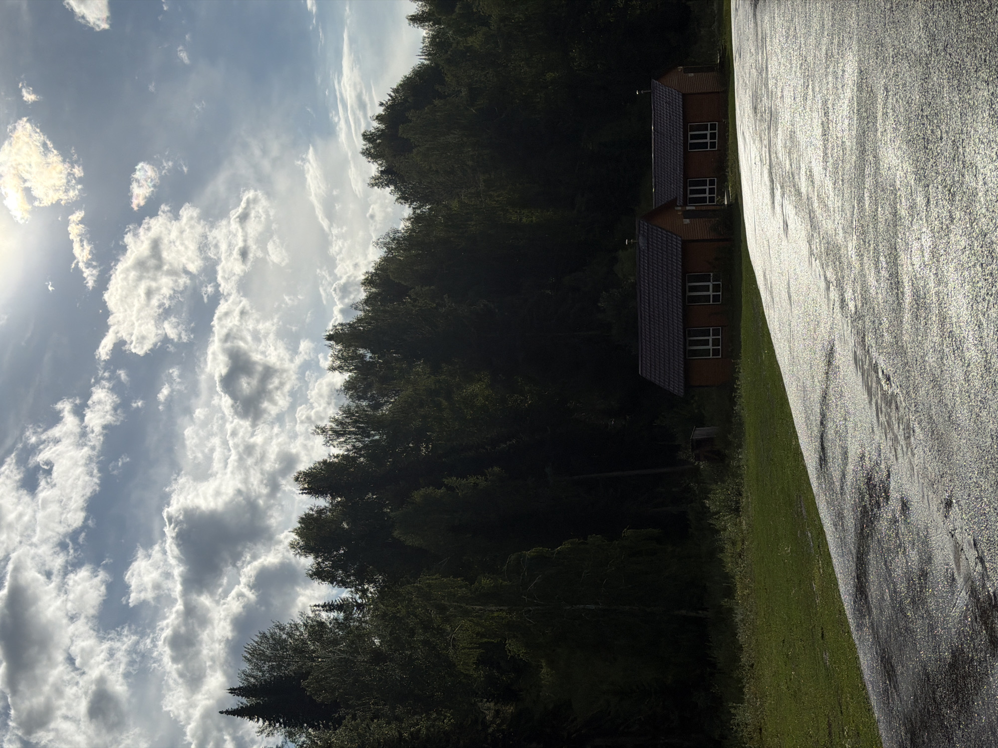 The image depicts a view from a wet, asphalt road towards two small, wooden houses with dark tiled roofs, nestled at the edge of a dense, dark green forest. The sky is filled with dramatic, sunlit clouds, suggesting a partially cloudy day. The foreground shows the glistening surface of the road, reflecting the light. The middle ground features a strip of green grass separating the road from the forest. The houses, identical in design with multiple windows, are positioned side-by-side. A small, weathered wooden shed is visible to the left of the houses, partially obscured by vegetation. The overall scene conveys a sense of rustic tranquility in a natural setting. The location is identified as a rural area within the Tarkhanka Rural District, Kazakhstan.