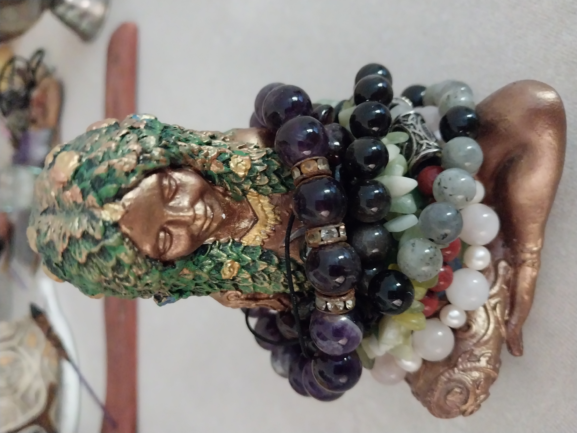 A close-up, eye-level shot shows a decorative figurine of a woman with a green and gold leafy headdress, adorned with multiple stacked bracelets around her lower body and base. The figurines face is downcast, with closed eyes and a gentle smile. The bracelets consist of various beads, including dark purple, black, white, light green, and some with metallic accents. The figurine appears to be made of a bronze-like material. The background is out of focus, showing hints of other objects and a pale surface, suggesting an indoor setting. There is no indication of location within the city of Dnipro, Ukraine.