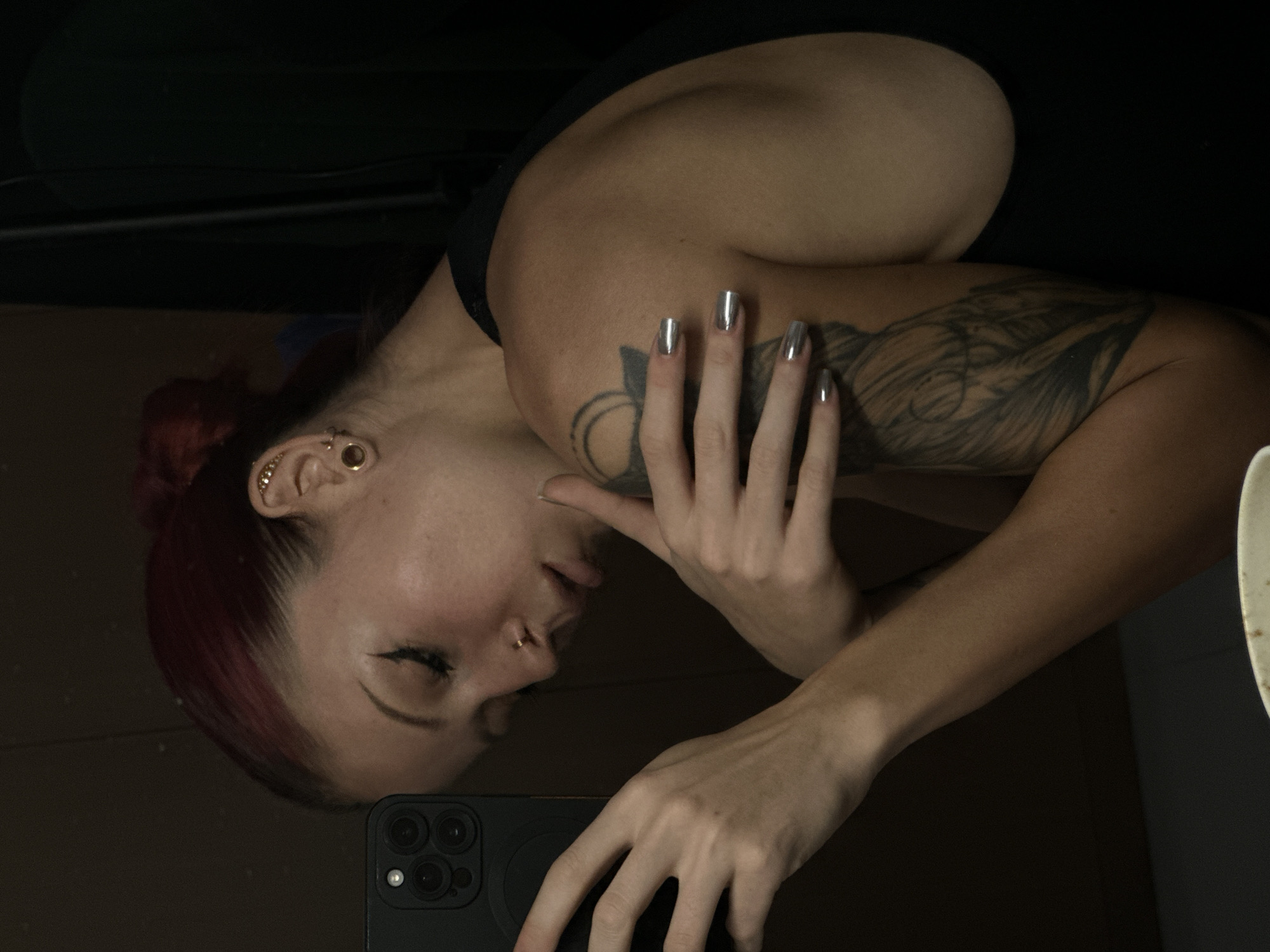 The image, oriented sideways, captures a close-up mirror selfie of a person with red or auburn hair, taken in what appears to be an indoor, dimly lit setting in North Kuta, Indonesia. The individual's face is seen in profile on the left side of the frame, with eyes closed or cast downwards, suggesting a contemplative or serene expression. They have several gold-toned piercings in their left ear and a small gold hoop piercing in their nose.
A prominent, large, dark tattoo featuring organic or possibly floral elements covers a significant portion of their left arm, extending onto the shoulder. Their fingernails are painted with a reflective, metallic silver polish. The person is holding a dark-colored smartphone, likely an iPhone given the distinctive triple-camera array, in their left hand, which is visible at the bottom of the frame. Their right hand rests near the phone, close to their face.
The background is largely dark and indistinct, with hints of a dark surface or wall behind. Part of a light-colored object, possibly a bowl or plate, is visible on the far right edge of the image. The person is wearing a dark top, with a strap visible over their shoulder. The overall lighting is low, creating a focused, intimate atmosphere. No visible text is present in the image.