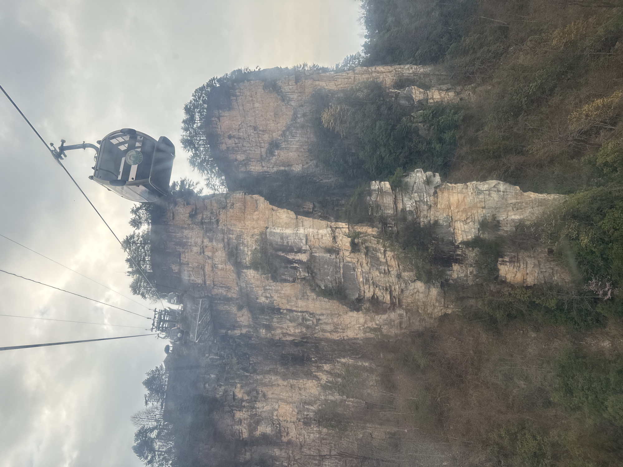 The image depicts a cable car suspended high above a dramatic mountainous landscape in Wulingyuan, Zhangjiajie National Forest Park, China, under an overcast sky. A single, light-colored cable car cabin, with darkened windows and a visible circular logo on its side, is positioned in the upper right quadrant, moving along a series of steel cables stretching across the scene. In the distance, towards the upper left, part of a cable car station, including a platform structure and at least one other cable car cabin, is visible atop a cliff face.
The environment is dominated by towering, sheer sandstone cliffs with vertical striations and varying earthy tones, including beige, grey, and dark brown, indicative of erosion and mineral staining. These cliffs are densely covered with vegetation, including coniferous trees on their summits and various deciduous and evergreen foliage clinging to ledges and slopes, appearing somewhat sparse and muted in color consistent with a non-peak growing season or high elevation. The sky is largely white and diffused, suggesting heavy cloud cover or atmospheric haze, which slightly obscures distant elements and softens the overall light. No people are discernibly visible within the cable car cabin or in the immediate landscape.