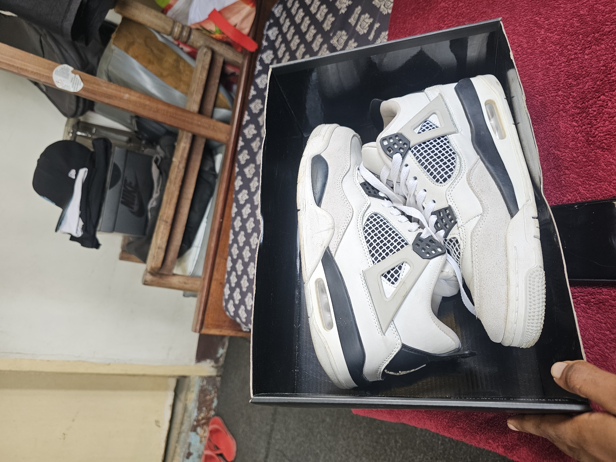 The image shows a pair of white and grey Air Jordan 4 sneakers placed inside a black shoe box. The box is positioned on top of a red textured surface, likely a rug or blanket. In the background, there is a wooden chair with a patterned cushion, and a portion of a wall painted in a light color. To the left of the chair, red flip-flops are visible on the ground. A hand with dark skin is partially visible on the left side of the frame, reaching towards the shoe box. The scene appears to be an indoor setting, potentially a residential area in Dar es Salaam, Tanzania, given the context provided. The lighting suggests it is daytime.