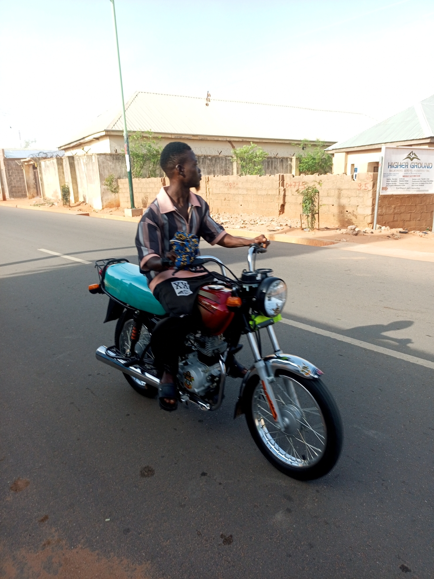 A man is riding a motorcycle on a paved road in Tempure, Nigeria. The motorcycle is red and silver with a turquoise seat. The man is wearing a striped shirt, black pants, and sandals. The background includes buildings, a fence, and a sign that reads "Higher Ground." The weather appears to be sunny.