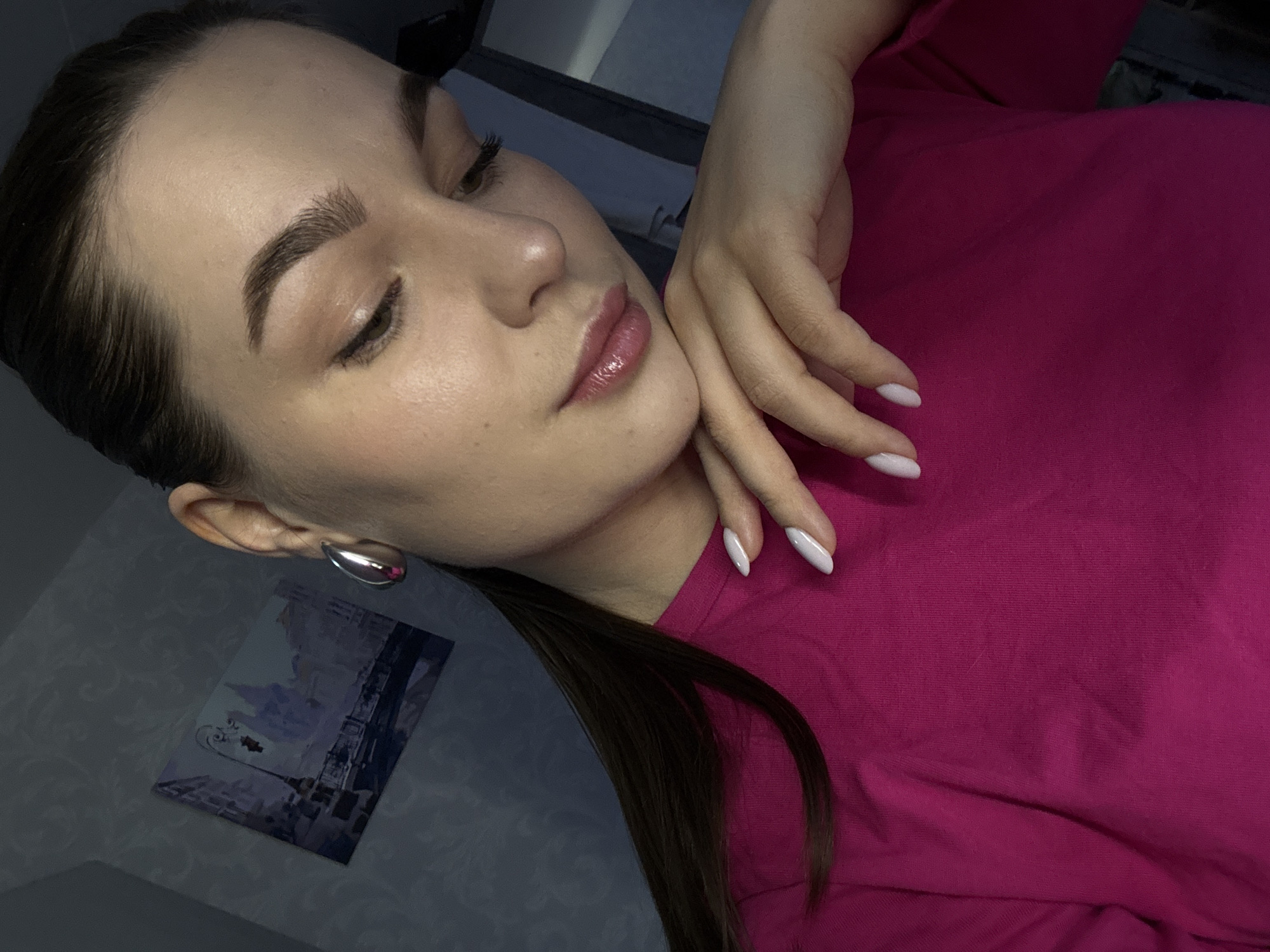 The image features a young woman reclining indoors, captured from an angled perspective. She appears to be lying on her side, with her head resting gently on her right hand, her eyes closed or partially closed in a serene expression. Her long, dark hair is styled in a low ponytail, visible over her left shoulder, with some strands falling forward.
She is wearing a vibrant magenta-colored top, possibly a t-shirt, which covers her torso. Her skin appears smooth and well-cared for, with subtle makeup accentuating her features: well-defined, full eyebrows and glossy, pinkish-nude lips. Her fingernails are manicured with a light, possibly white or very pale pink, polish and shaped in an almond style. A distinctive silver-toned, teardrop-shaped hoop earring adorns her visible left ear.
The background is dimly lit, suggesting an indoor setting with low ambient light, possibly evening. Behind her, a dark gray or blue wall is visible, on which a small framed picture hangs. The picture depicts a stylized cityscape, with what appears to be the Eiffel Tower, possibly hinting at Paris or a general travel theme.
The overall mood of the image is tranquil and contemplative. No visible text is present. This scene takes place in Chaikovsky, Russia.