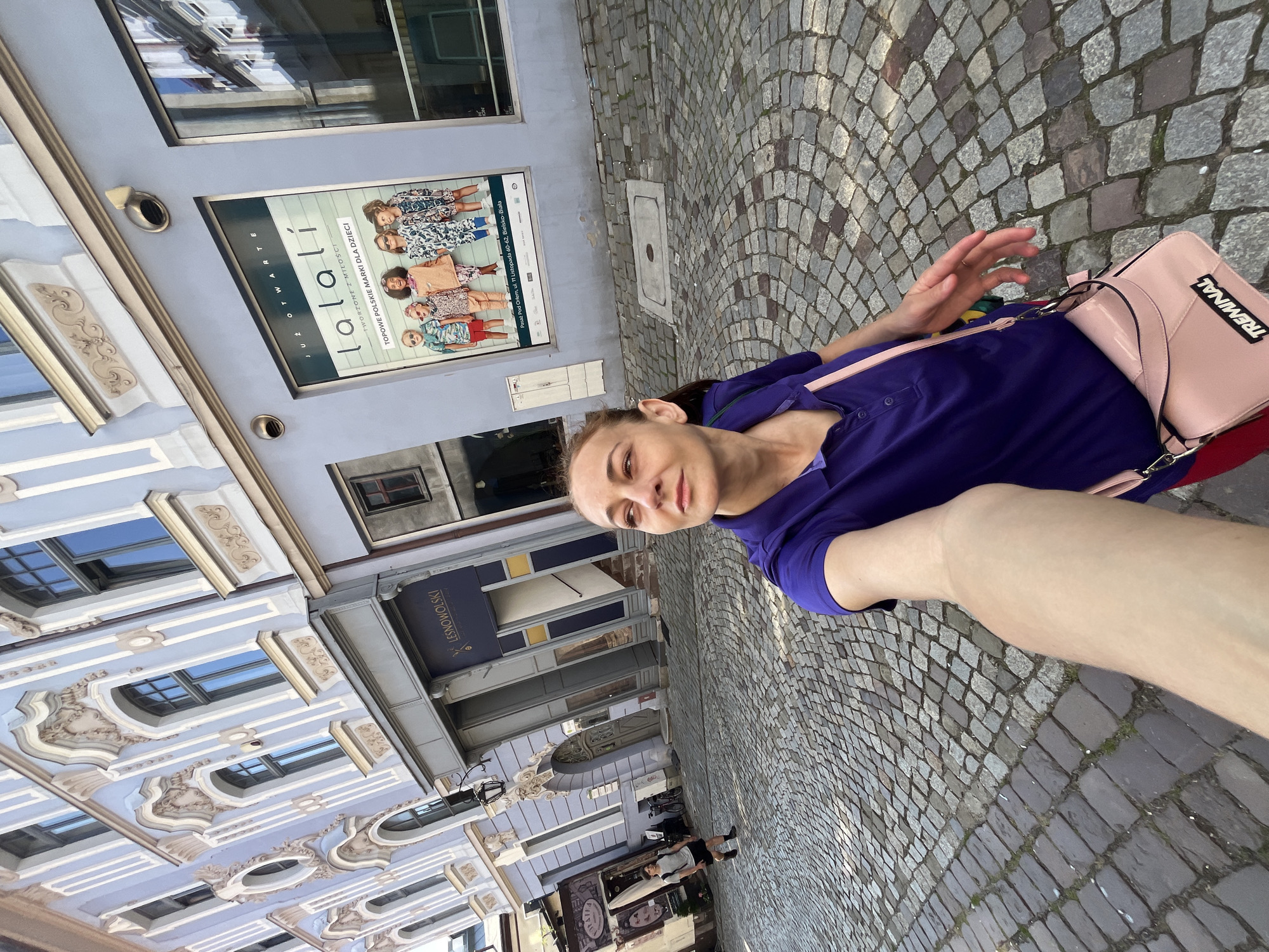 In this selfie, a woman with a light complexion and dark hair pulled back smiles at the camera, her face angled slightly. She is wearing a deep purple short-sleeved shirt and a pink crossbody bag with "TREMINAL" written on it in black. Her right hand is raised, with a ring on her index finger.
The woman is standing on a cobblestone street. Behind her, a light blue building features a poster advertising "lalalí" with images of children in colorful clothing. The text on the poster reads "JUŻ OTWARTE TWORZONE Z MIŁOŚCI TOPOWE POLSKIE MARKI DLA DZIECI" and mentions "Pasaż Pod Orłem, ul. 11 Listopada 60-62, Bielsko-Biała," indicating the location. Another building with ornate architectural details is visible to the left. The time of day appears to be daytime, with bright lighting suggesting fair weather. In the background, other people are walking on the street and a bicycle is parked. The overall scene depicts a casual outing in a European-style urban setting.