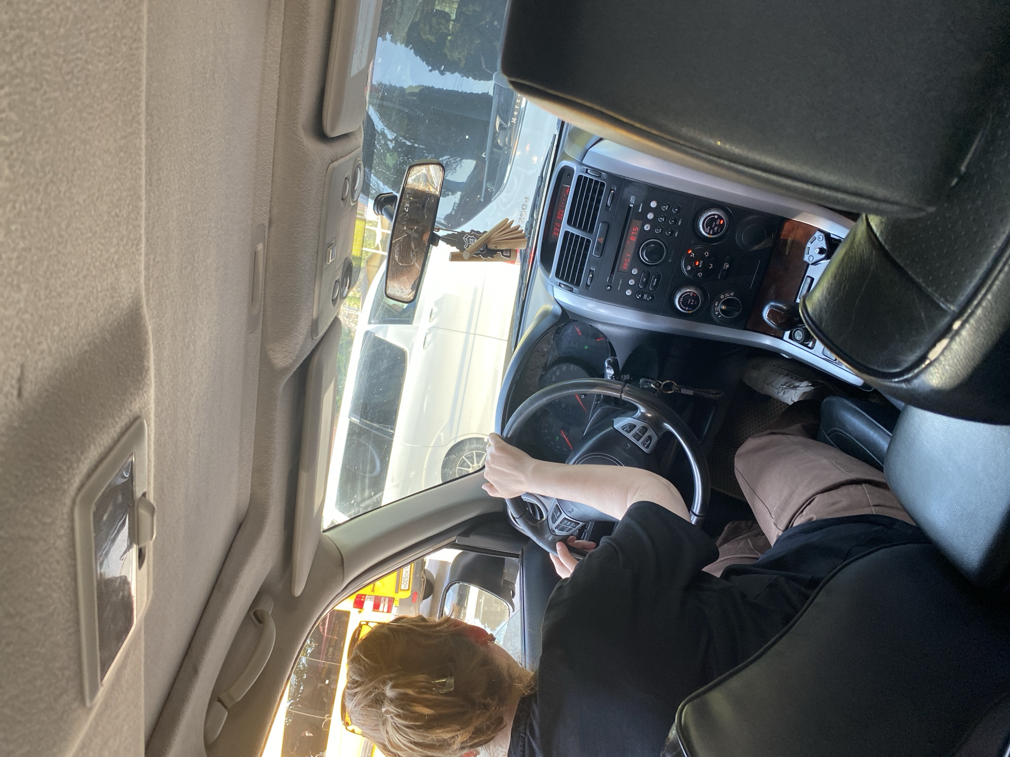 The image, captured from inside a car, shows the interior from the perspective of a passenger in the back seat, looking forward. The primary subject is the driver, a person with light brown hair pulled back in a bun, wearing a black t-shirt and brown pants. They are actively driving, with their hands on the steering wheel. The car's dashboard is visible, featuring a car stereo with a red digital display showing "23.1 2023," air vents, and various controls. A rearview mirror hangs from the windshield, reflecting the scene behind, and a car air freshener is suspended from it. Outside the car, other vehicles are visible, including a white SUV and a yellow vehicle. The environment appears to be a city street, with trees and sunlight suggesting an outdoor setting during the daytime. The lighting conditions indicate it is likely late afternoon due to the warm tones of the sunlight.