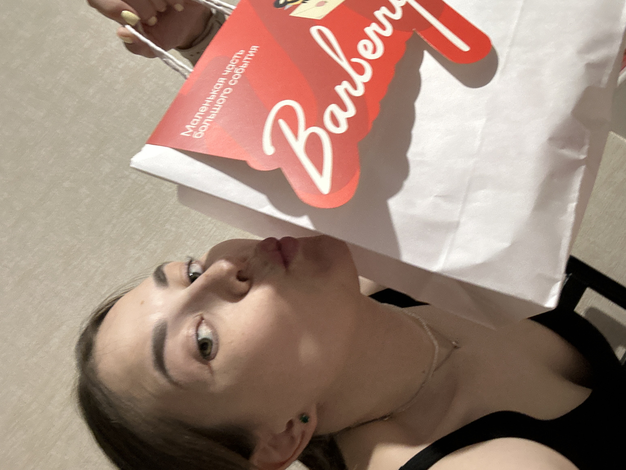 A young woman with dark hair and green eyes is leaning towards a white shopping bag with a red graphic that says "Barberry" in white cursive font. The bag also has Russian text that reads "Маленькая часть большого события" which translates to "A small part of a big event." She is puckering her lips as if to kiss the bag. The woman is wearing a black top and a delicate necklace. Her left earlobe has a small green earring. Another person's hand with yellow nail polish is holding the handle of the shopping bag. The background appears to be a neutral-colored wall. There is no visible indication that this scene is located in the city of Perm, Russia.