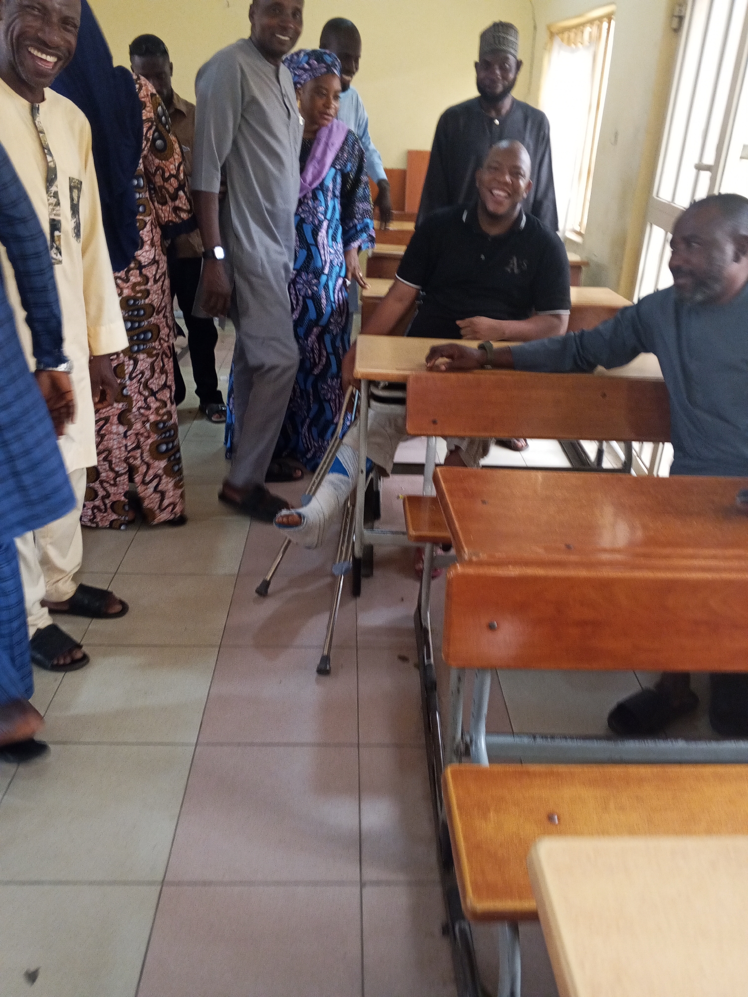 A group of people are gathered in a classroom setting in Lokoja, Nigeria. Several individuals are smiling, suggesting a positive or jovial atmosphere. One person, positioned in the foreground and slightly to the right, is seated at a wooden desk, looking towards the camera with a broad smile and a beard. Another man, standing behind him and to the left, is also smiling widely and is dressed in a black garment and a cap. To the left, a man in a light-colored, patterned garment is laughing heartily.
A significant detail is a person with a cast on their leg, using crutches to stand. This individual is wearing a blue and black patterned dress and a headscarf. The presence of desks and a classroom-like environment indicates this is likely an educational institution. The flooring appears to be tiled in beige and pinkish hues.
Other individuals in the background are partially visible, dressed in traditional African attire. The lighting suggests it is daytime. No text is visible in the image. The overall scene captures a moment of social interaction and perhaps a celebratory or welcoming gathering, despite the injury of one participant.