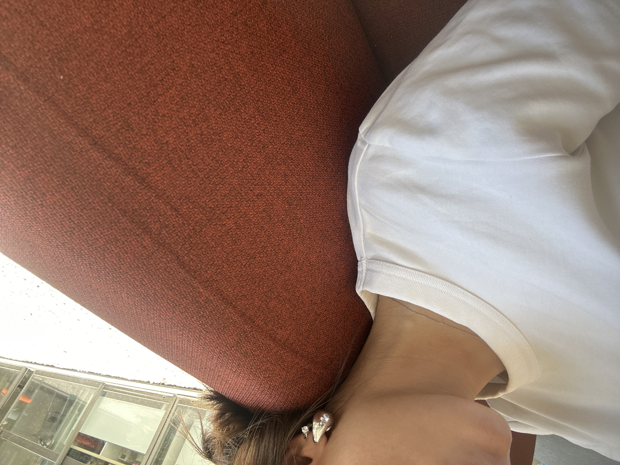 The image, rotated 90 degrees clockwise, captures a close-up, partial view of a person's upper body. The subject is seen from the neck down, or with their head tilted back, wearing a plain white t-shirt. Visible details include a bare neck adorned with a thin, silver-colored chain necklace, and the side of their head where brown hair meets the ear. The ear features two earrings: a prominent, sculptural or drop-shaped silver-colored piece and a smaller, sparkly stud.
A large portion of the frame is occupied by a textured, reddish-brown fabric surface, likely upholstery from a car seat, headliner, or a piece of furniture, positioned above the person's head. Below this surface, through a window, an outdoor scene is brightly illuminated by daylight. Architectural elements resembling glass panels and building structures are visible, suggesting an urban or commercial setting, possibly a drive-thru or office building. The scene depicts a static moment, with no specific activity discernible beyond the person seemingly resting or looking up within an enclosed space. There is no visible text in the image.