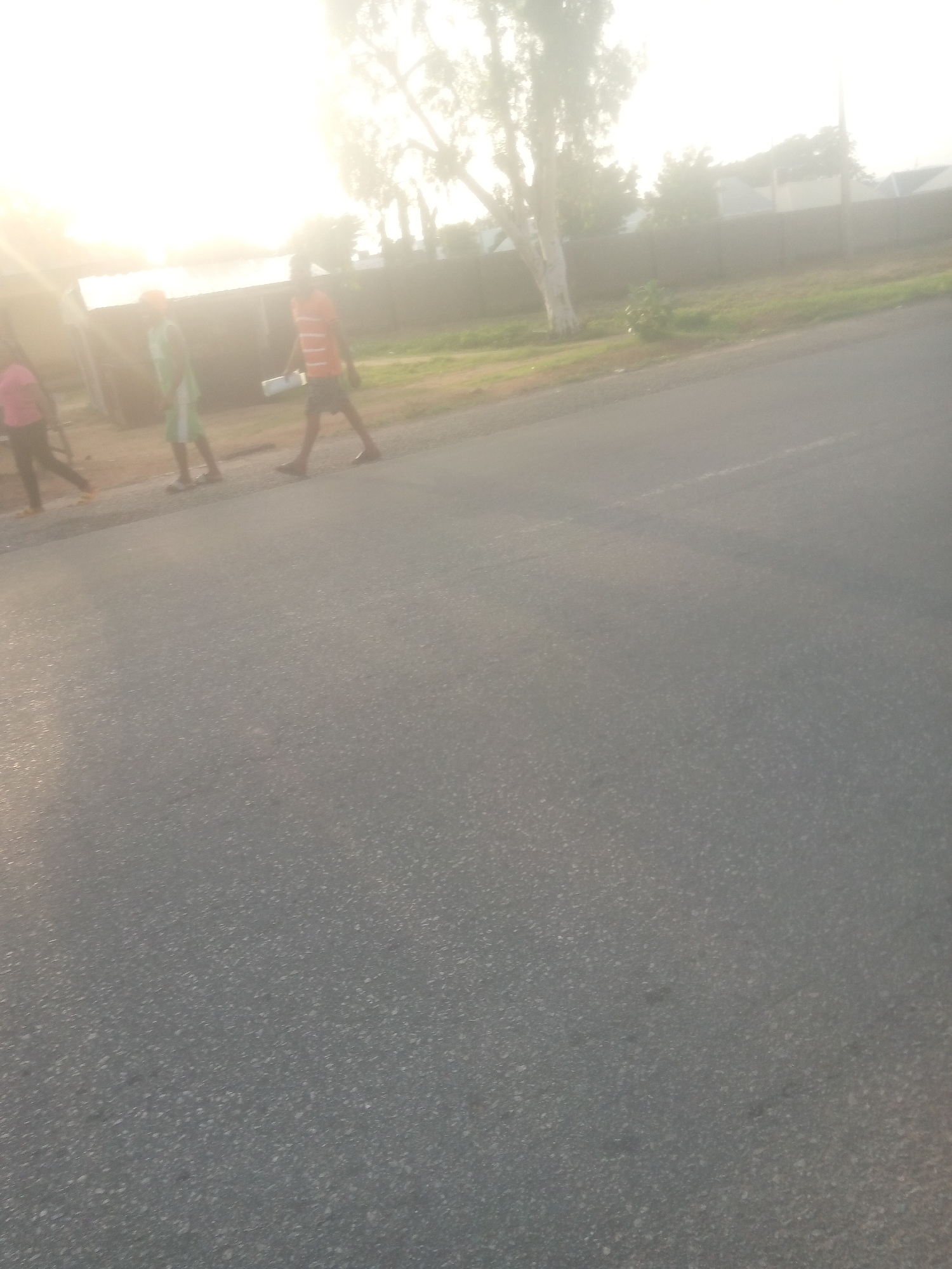 In Jalingo, Nigeria, on a paved road, three individuals are walking from left to right. The person on the far left is wearing a pink shirt and dark pants. Next to them, a person in a green shirt and white shorts walks. The third person, wearing an orange and white striped t-shirt and dark shorts, carries a white rectangular object. A small structure with a corrugated roof is visible behind them, partially obscured by bright sunlight. In the background, a chain-link fence separates the road from buildings and trees. The overall scene is illuminated by bright sunlight, creating a hazy effect.
