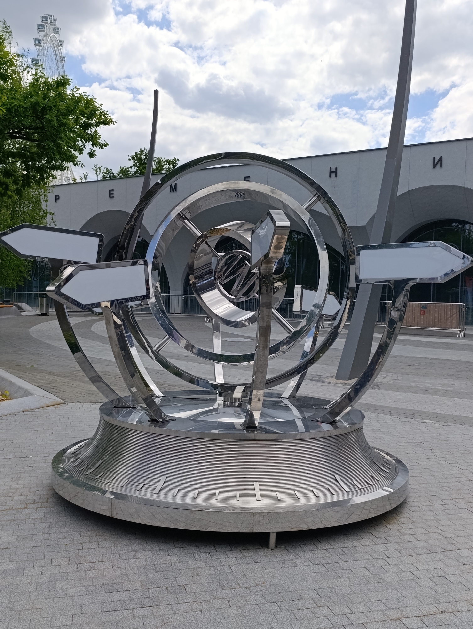 The image features a prominent, highly reflective chrome or polished metal sculpture, possibly an armillary sphere or sundial, set in an outdoor urban space. The intricate sculpture consists of multiple interconnected rings and a central sphere, all crafted from a polished, mirror-like material. Three blank, white, arrow-shaped signs are integrated into the sculpture, pointing in different directions. The entire piece rests on a wide, circular base with horizontal grooves and radial markings, indicative of a sundial.
In the background, a modern, light-colored building with large arched windows or entrances is visible. The Cyrillic letters "P", "E", "M", "H", and "И" are clearly displayed on its facade, forming part of the word "ВРЕМЕНИ" (VREMENI), meaning "Time." On the left, partially obscured by green foliage, a large white Ferris wheel is visible against a bright, cloudy sky. The ground is paved with light grey, rectangular cobblestones. The weather appears bright with significant cloud cover, suggesting daytime. The scene depicts a public plaza or park, located in Moscow, Russia, where the sculpture serves as a notable landmark.