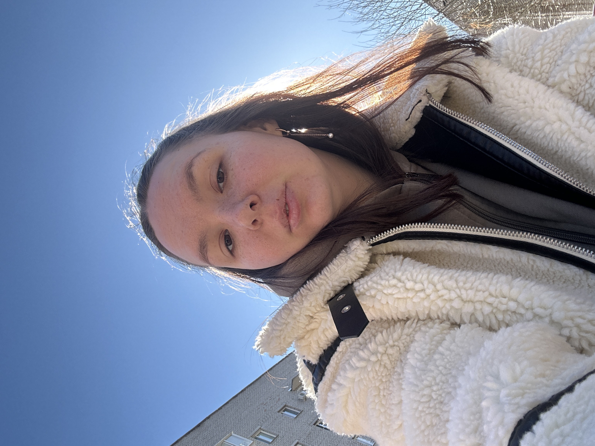 A young woman with long brown hair stands outdoors, looking slightly downwards into the camera. She wears a cream-colored fuzzy jacket with a black collar and a silver zipper. To her right, a building with a grayish facade and rectangular windows is visible. The sky above is clear and blue. Bare tree branches are partially visible in the upper right corner of the frame. The woman is wearing what appears to be a decorative earring with a chain and small pearl. The picture seems to be taken from a low angle, pointing upwards. Based on the building style and context, the location is likely in a residential area of Chaikovsky, Russia.