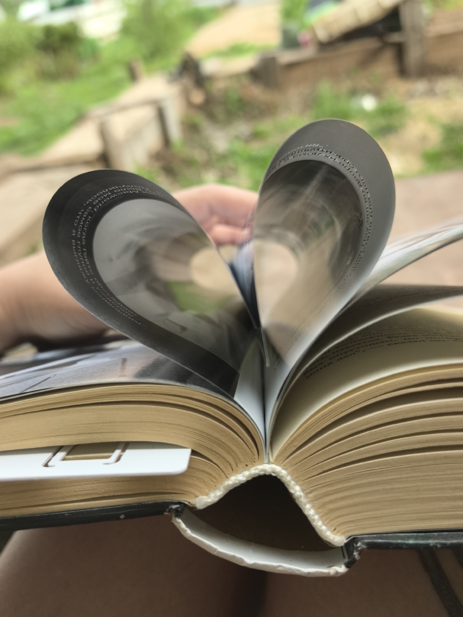 A close-up captures an open book with its pages bent into a heart shape. The book's pages are cream-colored with visible text, and a white bookmark is inserted. The book has a dark cover and its binding is visible. The background shows a blurry outdoor scene of greenery and wooden structures, suggesting a garden or park setting. A hand is faintly visible holding the pages.