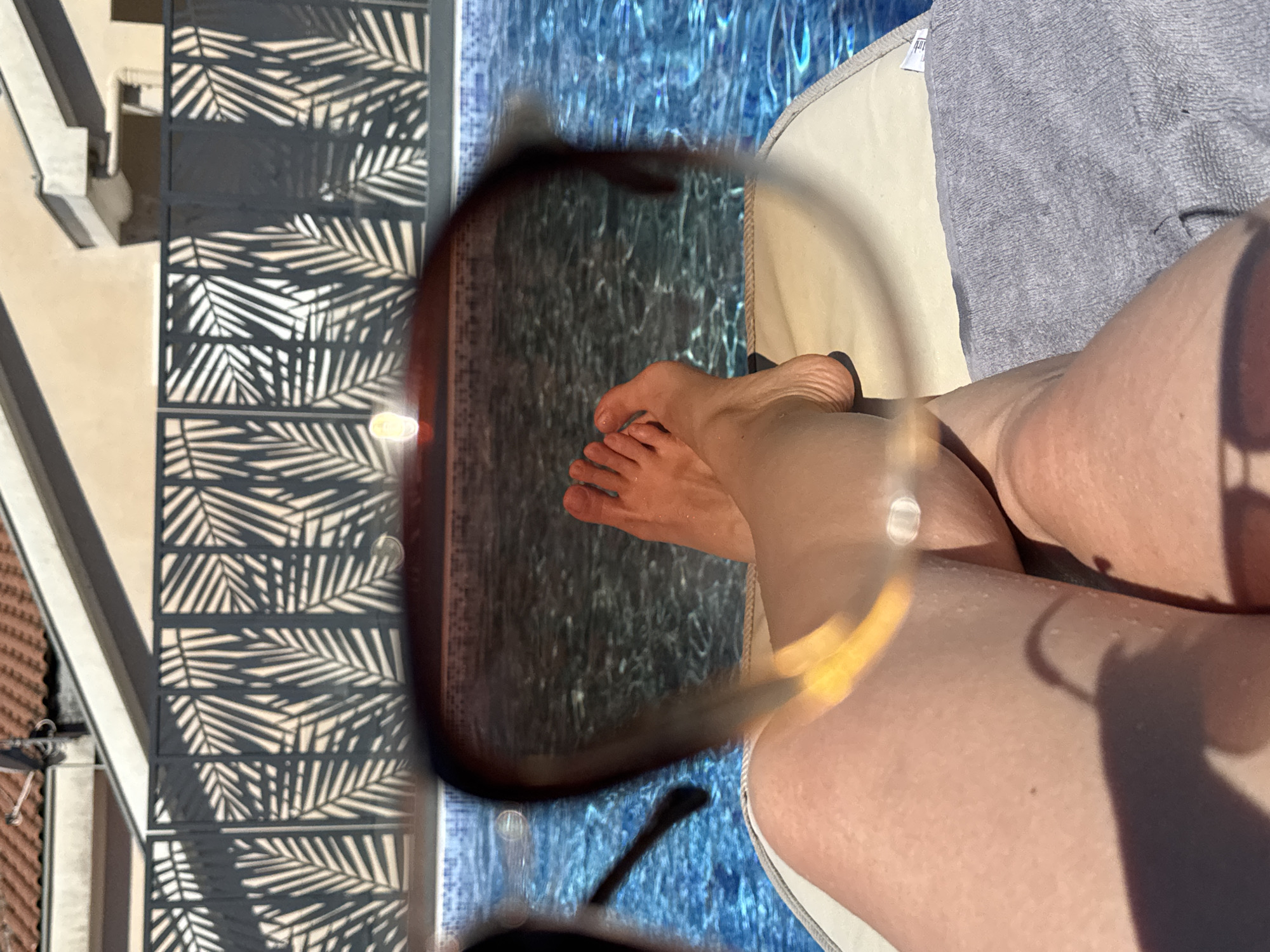 The image depicts a person lounging by a pool in Đenovići, Montenegro. The person's legs and feet are visible, and they are wearing sunglasses. The sunglasses reflect the water of the pool. A towel is underneath the leg and feet. In the background, there is a building with decorative black metal panels with leaf patterns.