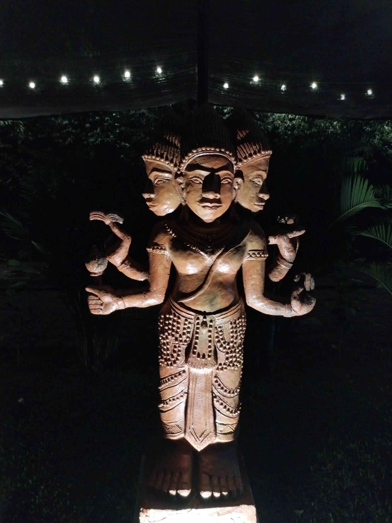 The image features a statue of a multi-faced, multi-armed figure, likely of religious significance, positioned outdoors under a dark sky with string lights in the background. The statue is illuminated from below, highlighting its intricate details and the textures of its material. The environment is surrounded by dark green foliage, suggesting a garden or outdoor setting. The location is identified as Bac Nha Trang Ward, Vietnam. No people are visible in the image, the sole subject being the statue itself.