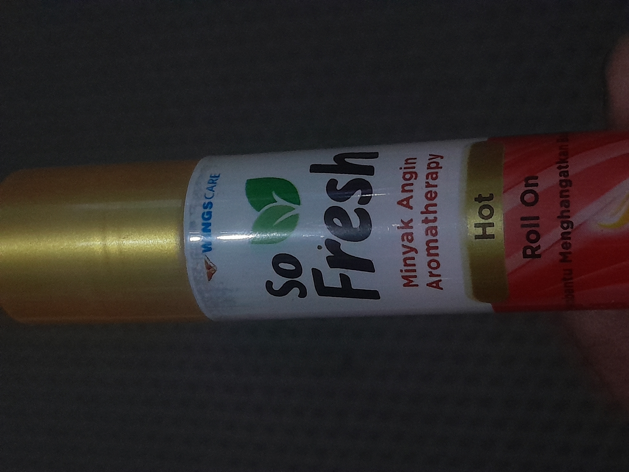 The image shows a close-up, slightly blurry, vertical shot of a "So Fresh" brand roll-on aromatherapy product against a dark, indistinct background. The product features a gold-colored cap. The label is white with blue and black text. Prominently displayed is the brand name "So Fresh" in large, bold, black lettering. Above this, in smaller blue text, is "WINGS CARE" and two green leaf icons. Below "So Fresh," in red text, is "Minyak Angin Aromatherapy." Further down, a yellow band with "Hot" in black text is visible, followed by "Roll On" in black text, and then "bantu Menghangatkan B" in smaller red text. The lighting is dim, with a slight glare on the bottle. There is no discernible location in Depok, Indonesia.