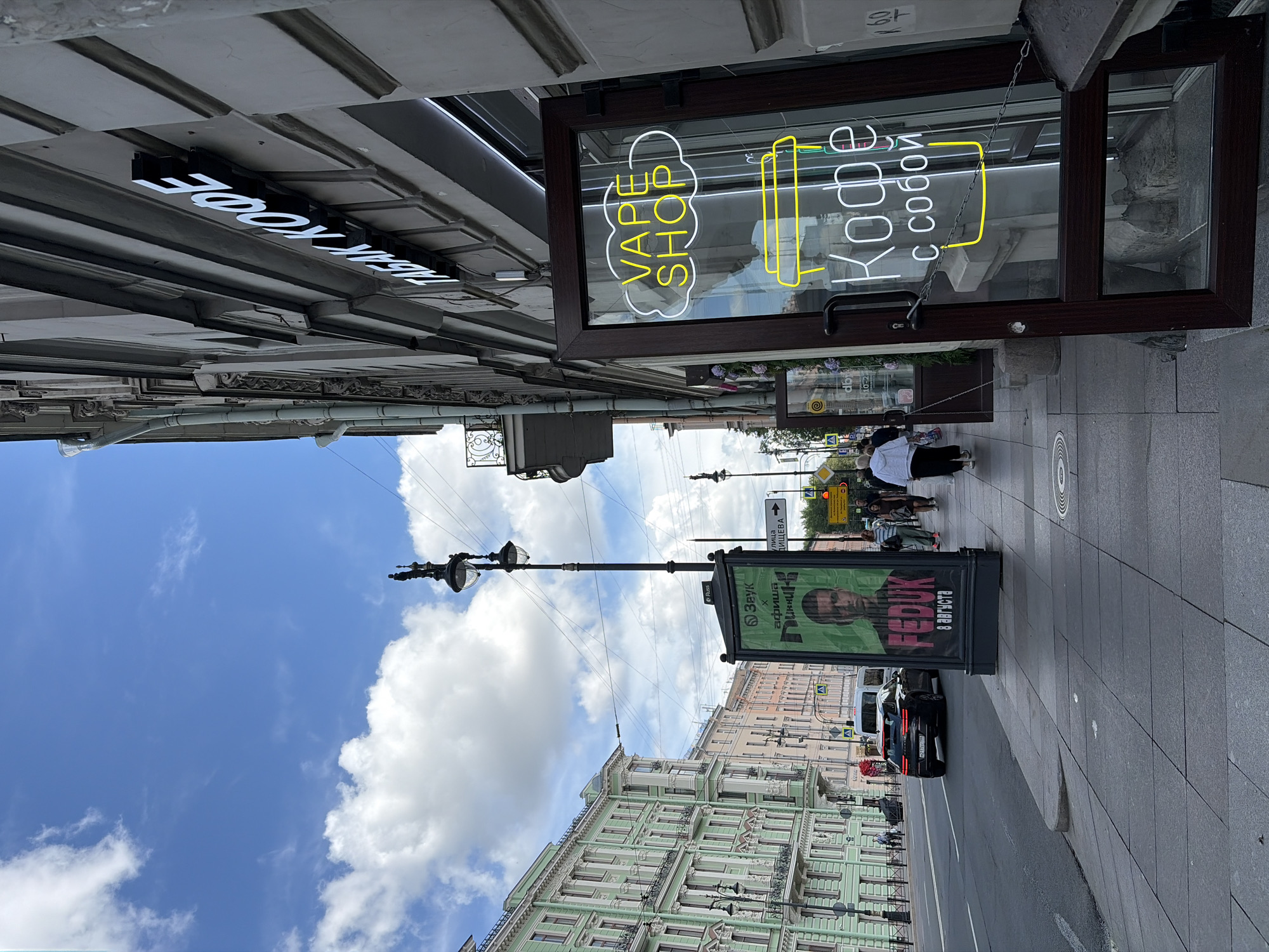 The image depicts a street scene in Saint Petersburg, Russia, likely on a sunny day with scattered clouds. The perspective is from a sidewalk, looking towards a building facade and the street.
On the right side of the frame, a building with a light-colored facade is prominent. A sign with white block letters on a dark background reads "ТАБАК КОФЕ", indicating a tobacco and coffee shop. Below this sign, a glass-door entrance to a shop is open, revealing a neon sign inside. The neon sign glows yellow and white, forming the words "VAPE SHOP" within a cloud shape, and below that, a yellow outline of a coffee cup and the words "Кофе с собой" (Coffee to go) in white neon. The door frame is dark brown, and a chain is looped across the opening.
To the left of the building, a sidewalk runs parallel to the street. Several pedestrians are walking along the sidewalk. One person in the foreground wears a white jacket and dark pants. Further down the sidewalk, more people are visible.
In the middle ground, a large advertising pillar stands on the sidewalk, displaying a poster with a portrait of a man and Russian text. A white van is parked or moving on the street near the pillar, and a black car is driving away from the viewer.
Further down the street, a line of multi-story buildings with ornate architecture lines the left side of the frame. These buildings are painted in various pastel colors, including a prominent pale green building with numerous windows and balconies. Streetlights and traffic signals are visible along the street and sidewalk. The sky above is blue with large, fluffy white clouds.