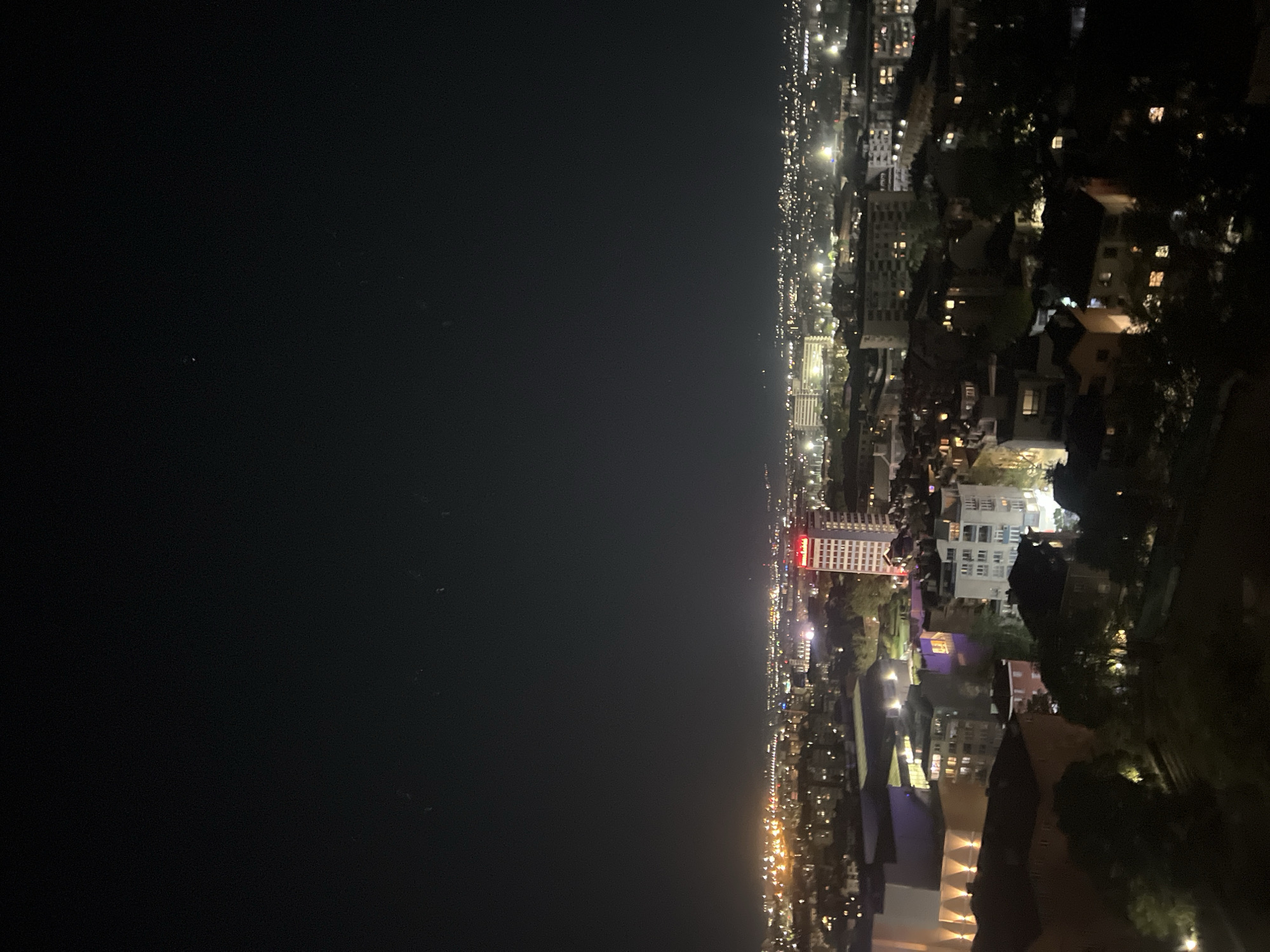 The image is a nighttime view of Plovdiv, Bulgaria, from an elevated position. The city lights illuminate the buildings and streets, contrasting with the dark sky. The left side of the image appears to be water. The scene shows a residential area with scattered trees and shrubs.
