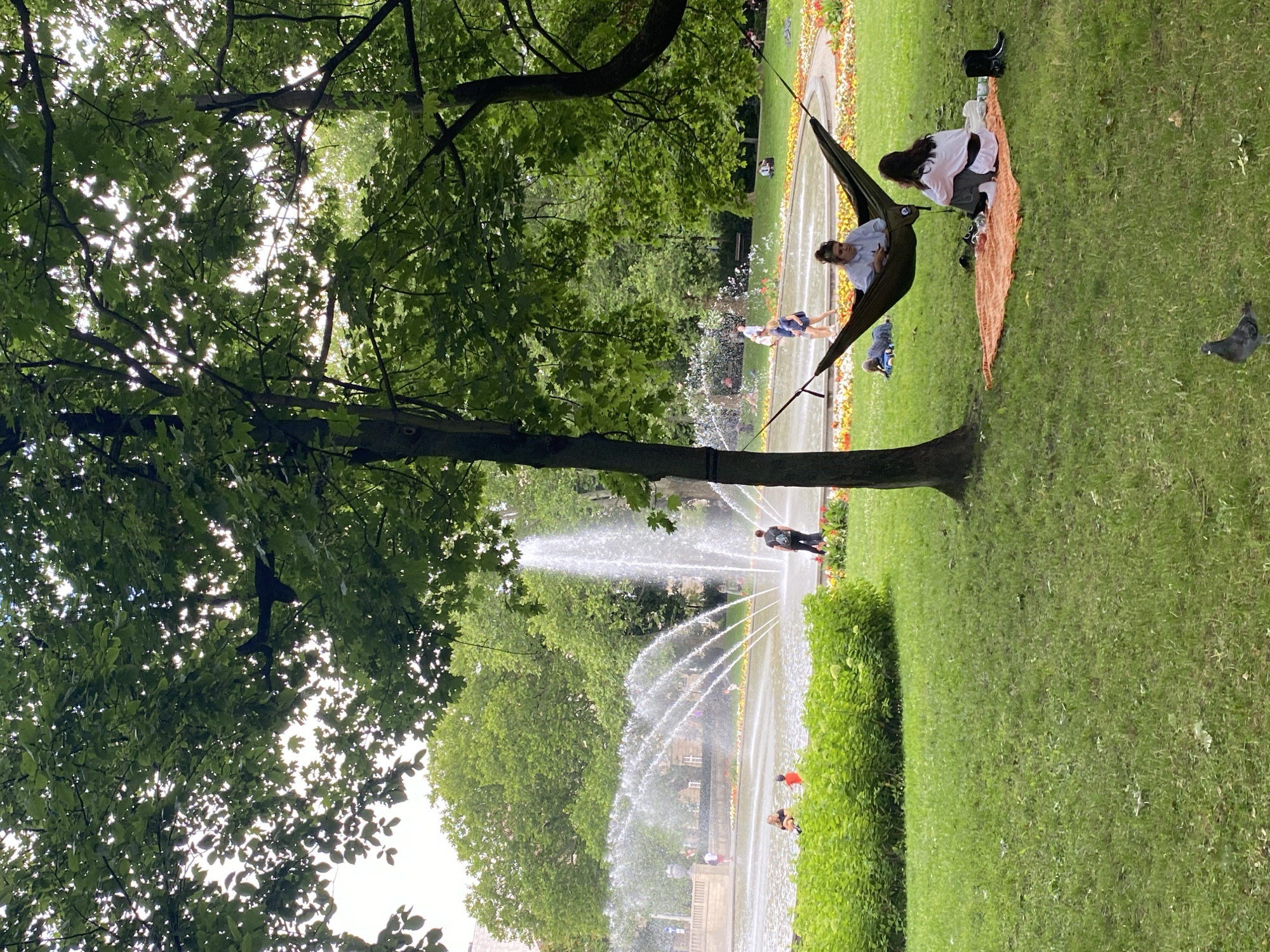 The image depicts a park scene in Poznan, Poland, with a large fountain in the background. In the foreground, two women are relaxing on a picnic blanket under a hammock hung between two trees. One woman is seated in the hammock, looking at her phone, while the other is sitting on the blanket. Several other people are visible in the park, some near the fountain and others walking in the distance. The park is covered in green grass and surrounded by trees and flower beds. A pigeon is also present on the grass in the lower part of the image.