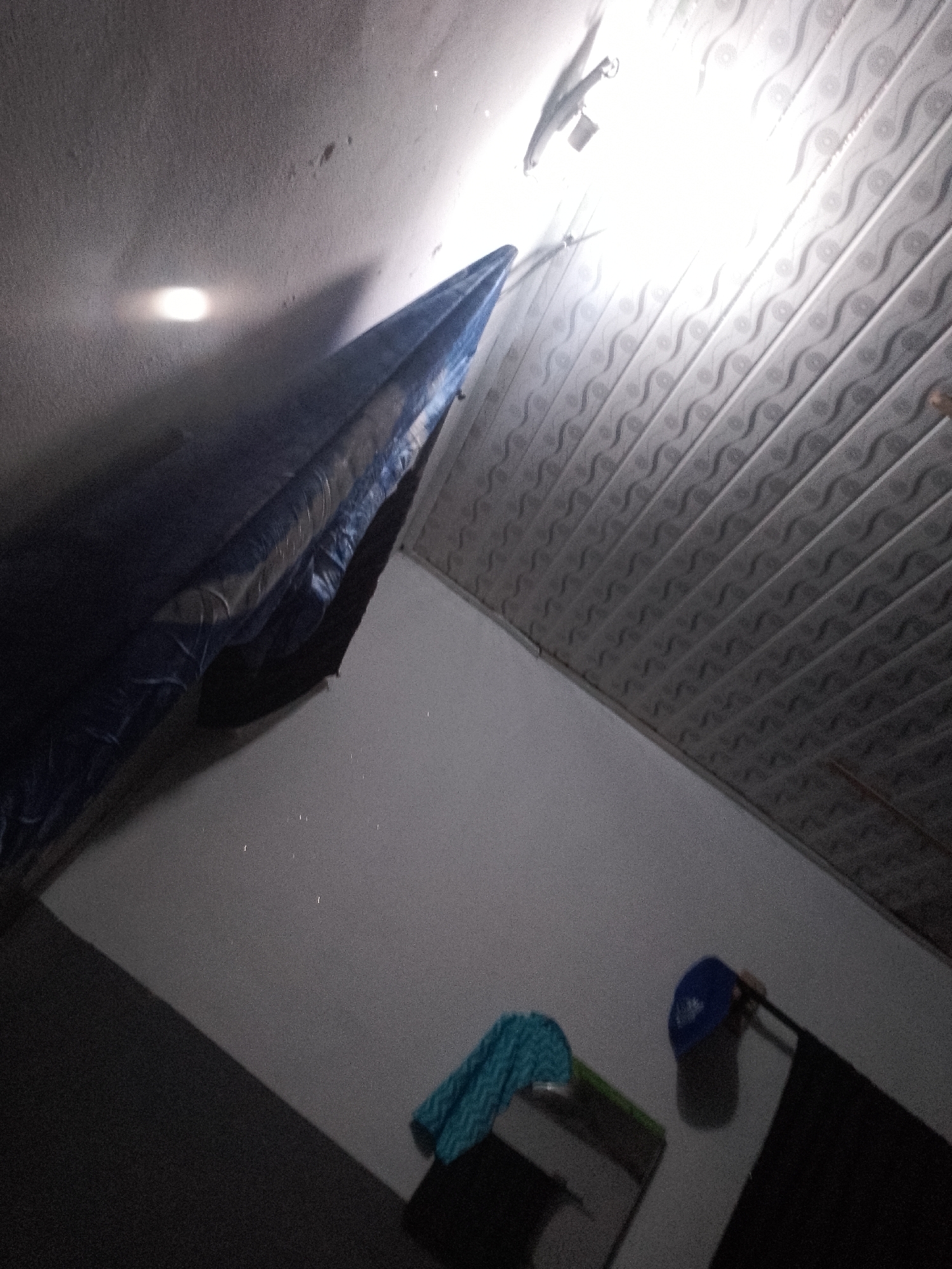 A dimly lit interior scene shows a ceiling with a patterned texture and a bright overhead light. Below the ceiling, a blue tarp is draped across an unseen object, casting a shadow on the wall. To the right, a blue baseball cap rests on a dark surface, possibly a shelf or table. A folded blue and white patterned cloth is also visible nearby. The scene appears to be an indoor storage area or unfinished space.