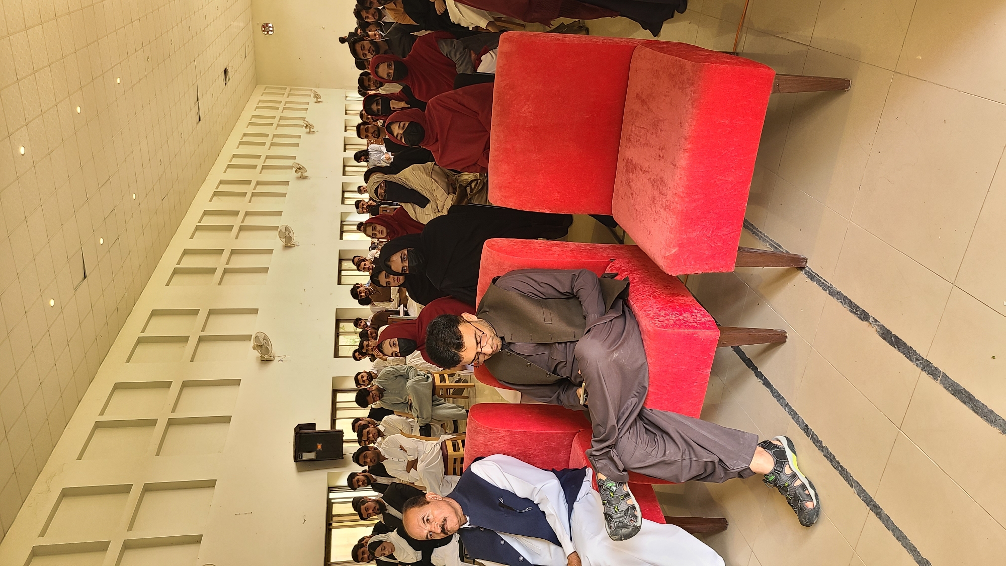 A medium shot, rotated counter-clockwise, shows a gathering of people in what appears to be a large indoor hall, potentially within the Quetta City Tehsil, Pakistan. Most attendees are seated on red upholstered armless chairs arranged in rows. The audience has both men and women. Some women wear headscarves or veils. Men are dressed in a variety of traditional Pakistani attire, including shalwar kameez and vests.
The hall's walls are white, with rectangular recessed decorations running horizontally near the ceiling. Small ceiling fans are spaced along the wall. The floor is tiled. The lighting seems bright and even.