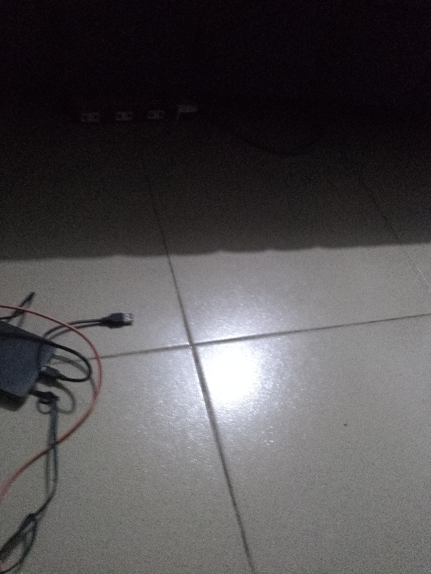 A close-up, low-light shot shows a black power brick or adapter with red and black wires connected to it, lying on a tiled floor. The adapter is dark and difficult to make out the details of, but the wires are clearly visible, one red and one black, looping around the adapter. A USB cable is also attached to the adapter, with its connector visible at the end. The floor is made of light-colored tiles with visible grout lines. A bright reflection, possibly from a light source off-camera, shines on the tiles in the middle of the image. The background is dark and out of focus, appearing to be a piece of furniture or wall.
