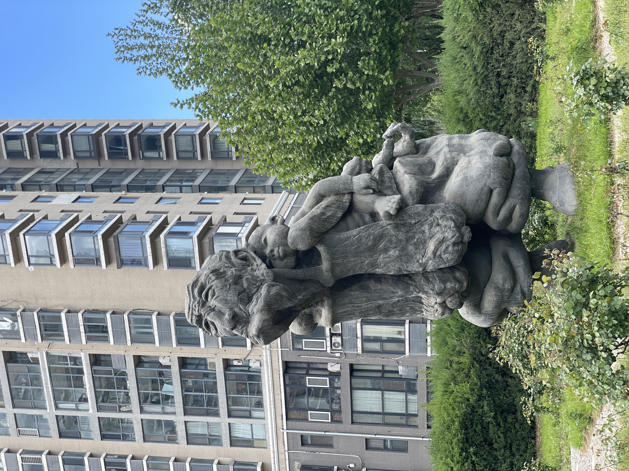 The image depicts a stone sculpture located outdoors in Changping District, China. The sculpture portrays a figure with what appears to be a bearded head and multiple intertwined bodies or limbs above it. The sculpture is positioned on a grassy area, surrounded by greenery, including bushes and trees. In the background, there are multiple buildings, one with a repeating pattern of windows and another with more modern windows. The sky is blue.