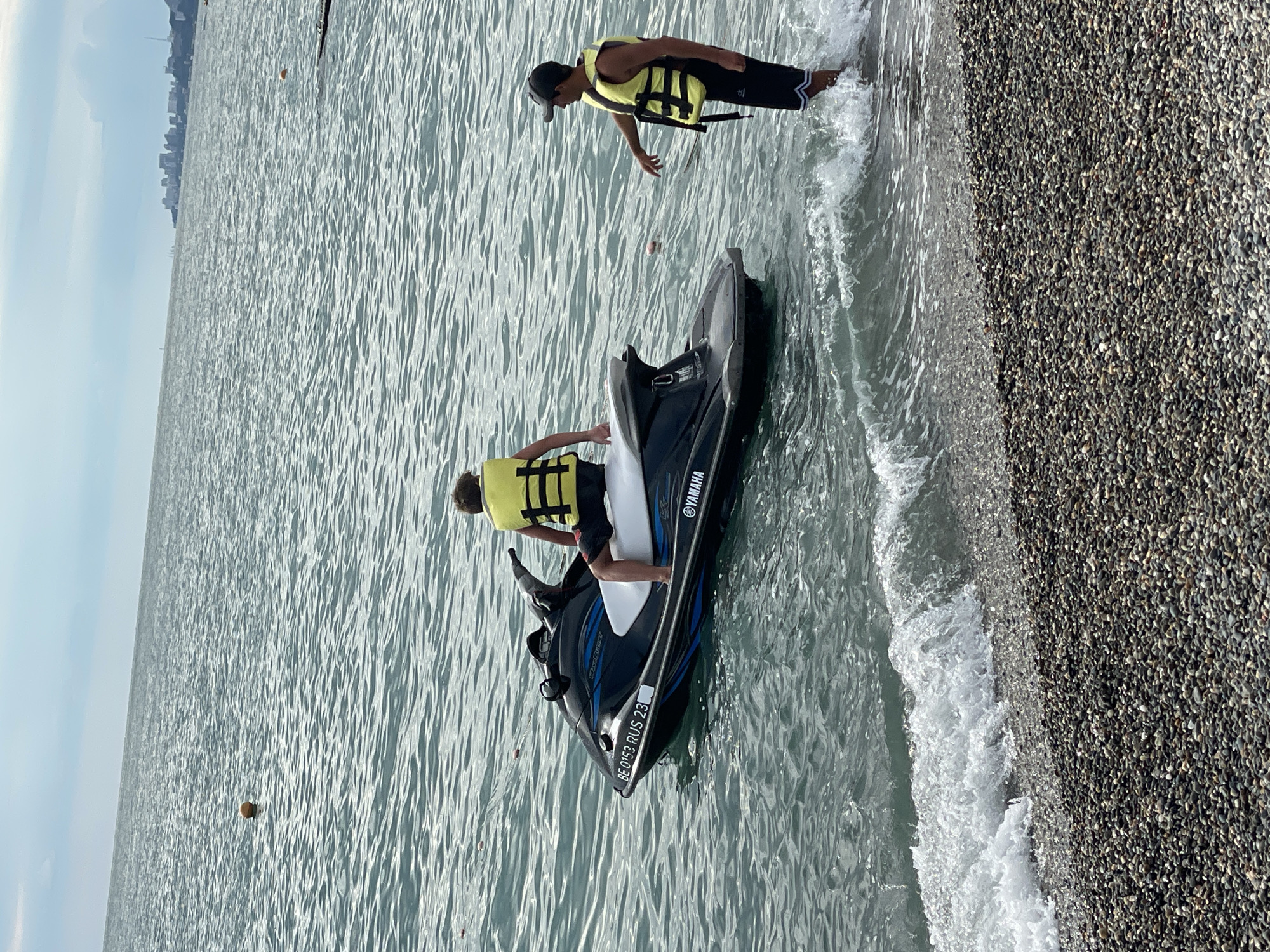 Two individuals are present near a jet ski in the Black Sea off the coast of Sochi, Russia. One person, a young male wearing a yellow life jacket and black and red patterned shorts, is seated on the jet ski. The jet ski is black with blue accents and white seating, bearing the "Yamaha" logo and registration "BE 0153 RUS 23". The second individual, an adult male wearing a black baseball cap, a yellow life jacket, and black shorts with white stripes, is standing in the water near the jet ski, appearing to interact with it. The water is calm with small waves lapping at the pebbled shoreline in the foreground. In the distance, a cityscape with buildings and structures is visible on the horizon, characteristic of Sochi. The sky is overcast.
