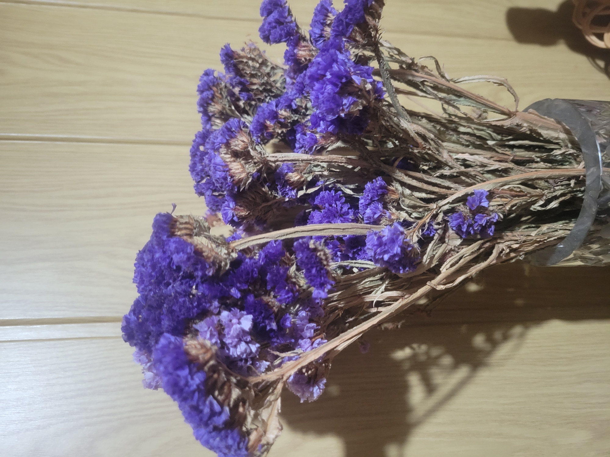 In Belaazyorsk, Belarus, a bouquet of dried statice flowers, predominantly a vibrant purple, is arranged in a clear glass vase. The flowers, characterized by their small, clustered blooms and thin, dried stems, are positioned in the foreground. Behind the bouquet, a plain, light-colored wooden wall serves as the backdrop. The lighting creates soft shadows on the wall, suggesting an indoor setting, possibly a home or a shop. The overall scene is still and quiet, with no people or active events depicted.