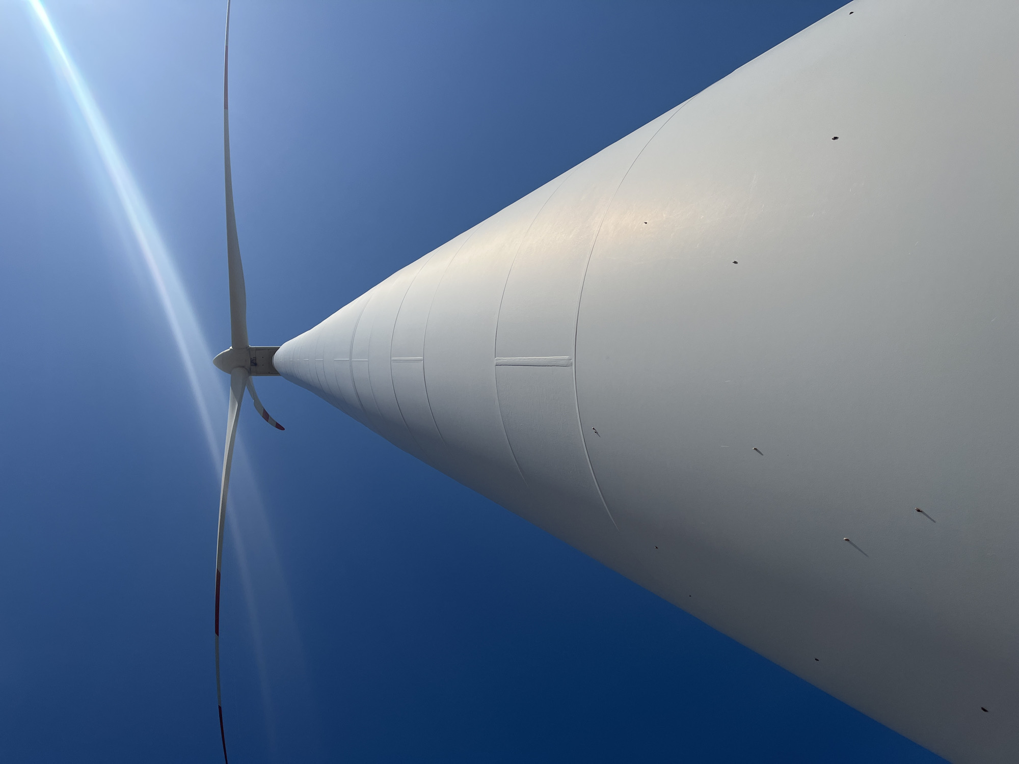 The image shows a close-up, low-angle view of a white wind turbine tower against a clear blue sky. The tower is segmented and tapers towards the top, where a large rotor with two blades is visible. The blades are white with red markings at the tips. A lens flare from the sun is prominent in the upper right quadrant of the image. The scene appears to be outdoors with no other objects or people visible. The location is identified as Rignano Garganico, Italy.