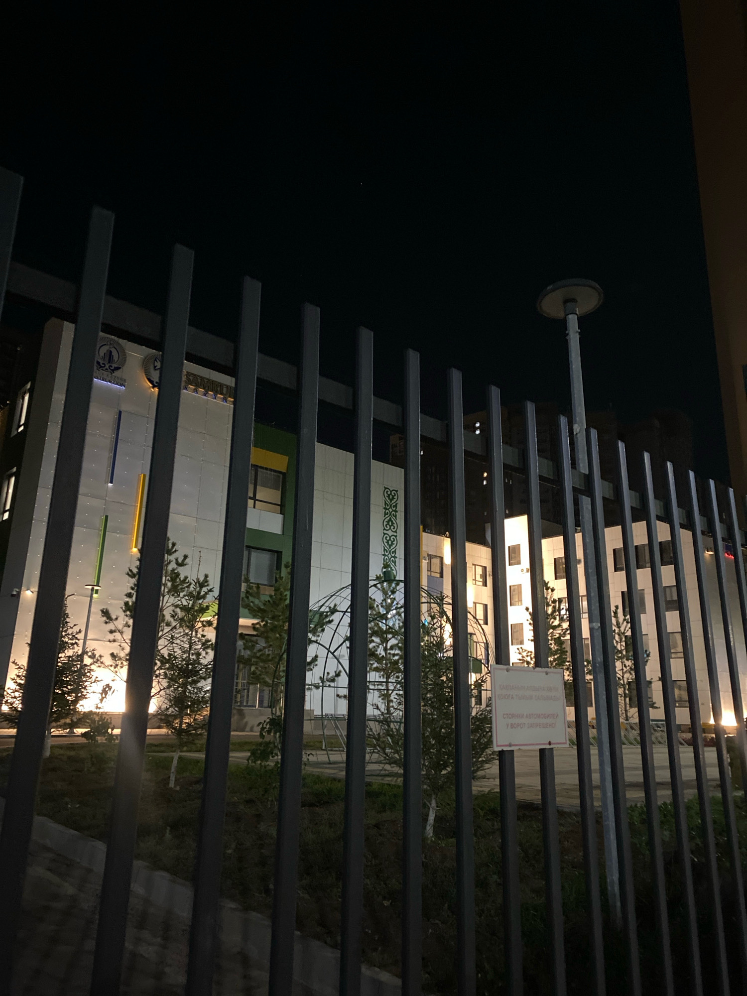 An exterior nighttime, eye-level shot captures a modern building facade behind a dark metal fence in Astana, Kazakhstan. The building, illuminated from within, features a white exterior with vertical green and yellow accents and a patterned design. A circular light fixture on a pole is visible above the fence, and a sign with Russian text is attached to the fence. Small trees are planted in front of the building. The sky is dark with a few faint stars.