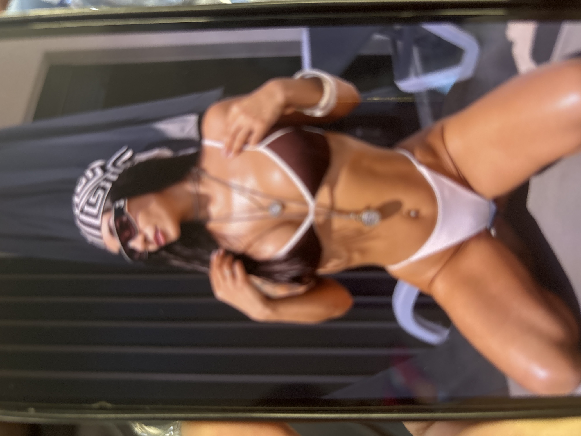 An image depicts a photograph displayed on a handheld digital screen, partially obscured by the fingers and thumb of a person holding it. The photograph features a young adult female in an outdoor setting in Blagoevgrad, Bulgaria. She is kneeling on a light-colored ground surface, facing slightly to the viewer's left with her head turned to the left. She wears a dark brown triangular bikini top with thin white trim, white high-cut bikini bottoms, dark wraparound sunglasses, and a black and white geometric patterned headscarf.
Her skin is tanned and appears to have a prominent sheen, suggesting the presence of oil, water, or sweat. She wears multiple silver-colored necklaces with circular pendants, one of which reaches below her navel, where a silver belly button piercing is visible. A wide, light-colored bracelet adorns her right wrist. Her left hand is raised, lightly grasping her dark, long hair near her shoulder, while her right hand touches the strap of her bikini top on her left shoulder.
The background consists primarily of a dark, vertically slatted structure, possibly a wall or fence. In the lower right background, a white outdoor lounge chair is partially visible. The overall lighting suggests a bright, sunny environment.