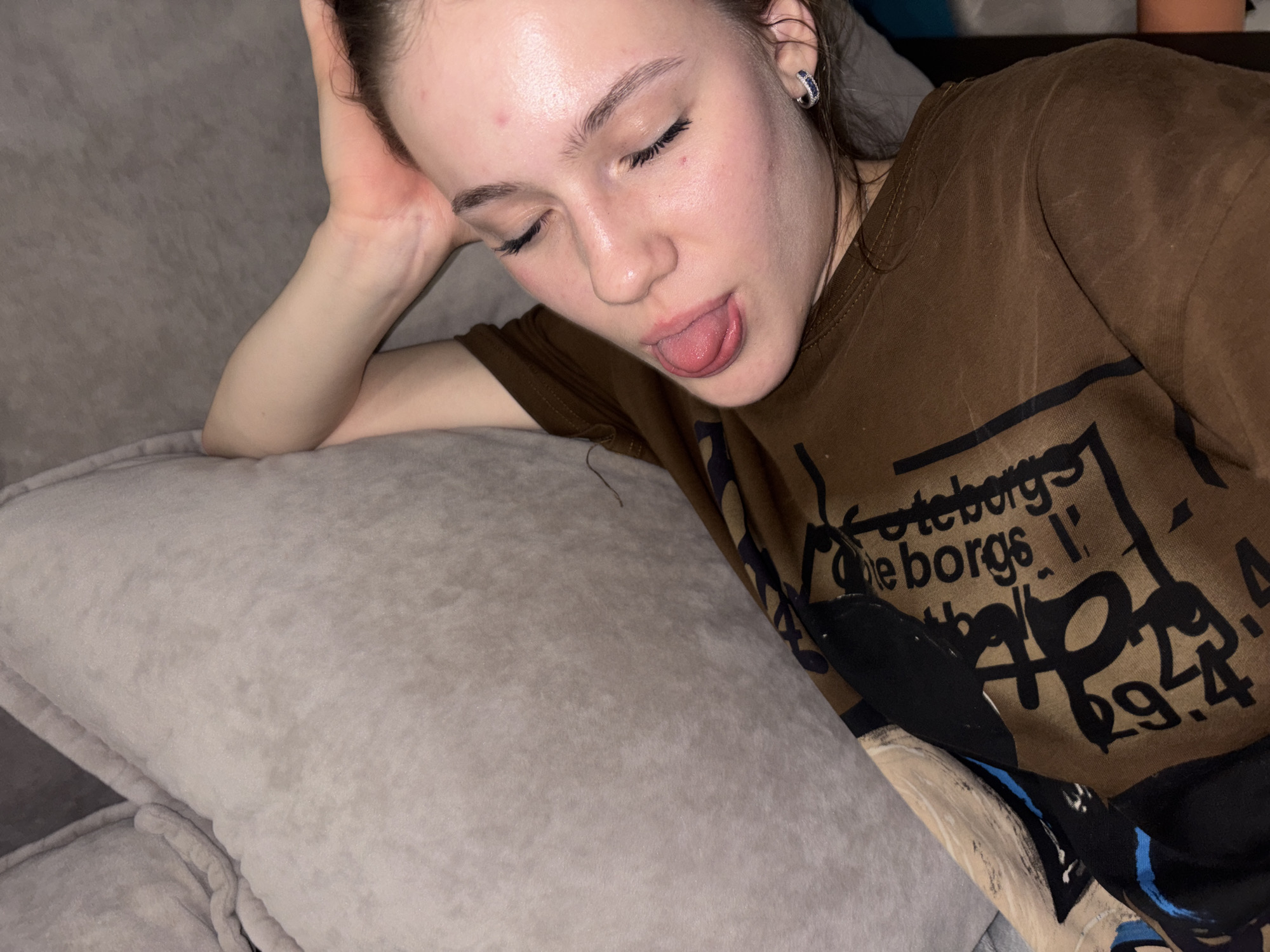 A young woman with her eyes closed and tongue sticking out is lying on a couch. She is wearing a brown t-shirt with a graphic print. A gray pillow is behind her head. The lighting is dim. The image is a close-up shot, and no specific details of Vaganovo, Russia are visible.