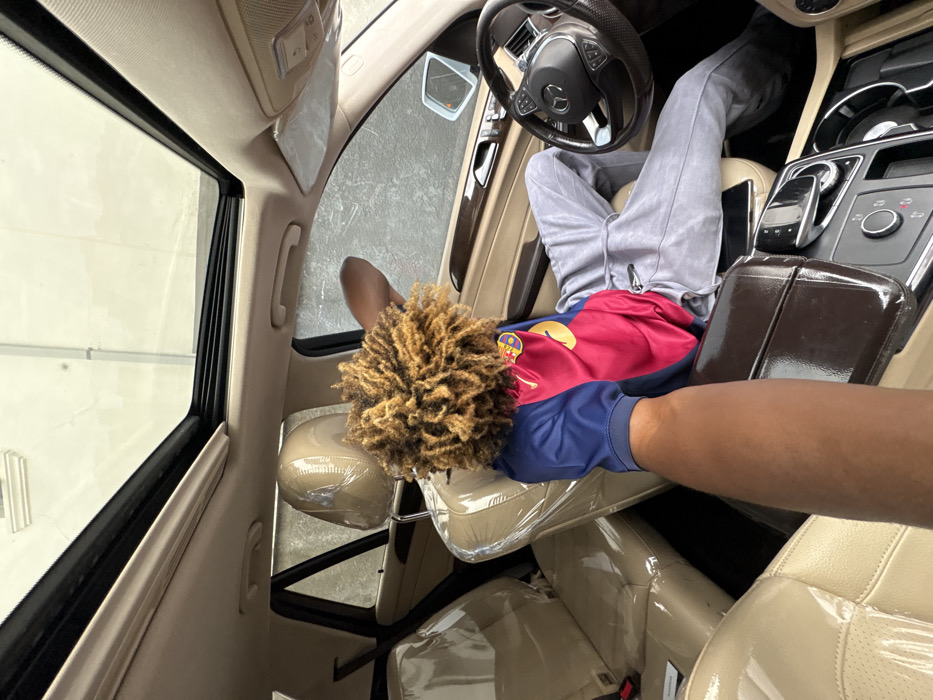 The scene is set inside a beige-interiored Mercedes-Benz SUV, located in Igboefon, Nigeria. A person with tightly coiled, light brown hair is seated in the front passenger seat, facing away from the camera. They wear a short-sleeved shirt, predominantly blue with a large red section on the back and a yellow emblem, paired with grey sweatpants. Their right arm is extended, resting on the upper interior door frame as they look out the passenger window. The rear passenger seats and headrests are covered in clear protective plastic. In the foreground, a dark-skinned arm extends from the bottom left, positioned towards the vehicle's center console, where a smartphone rests near the gear selector. Through the panoramic sunroof, a light-colored building structure or ceiling is visible. Outside the passenger window, a building with textured, light-colored walls is observed.