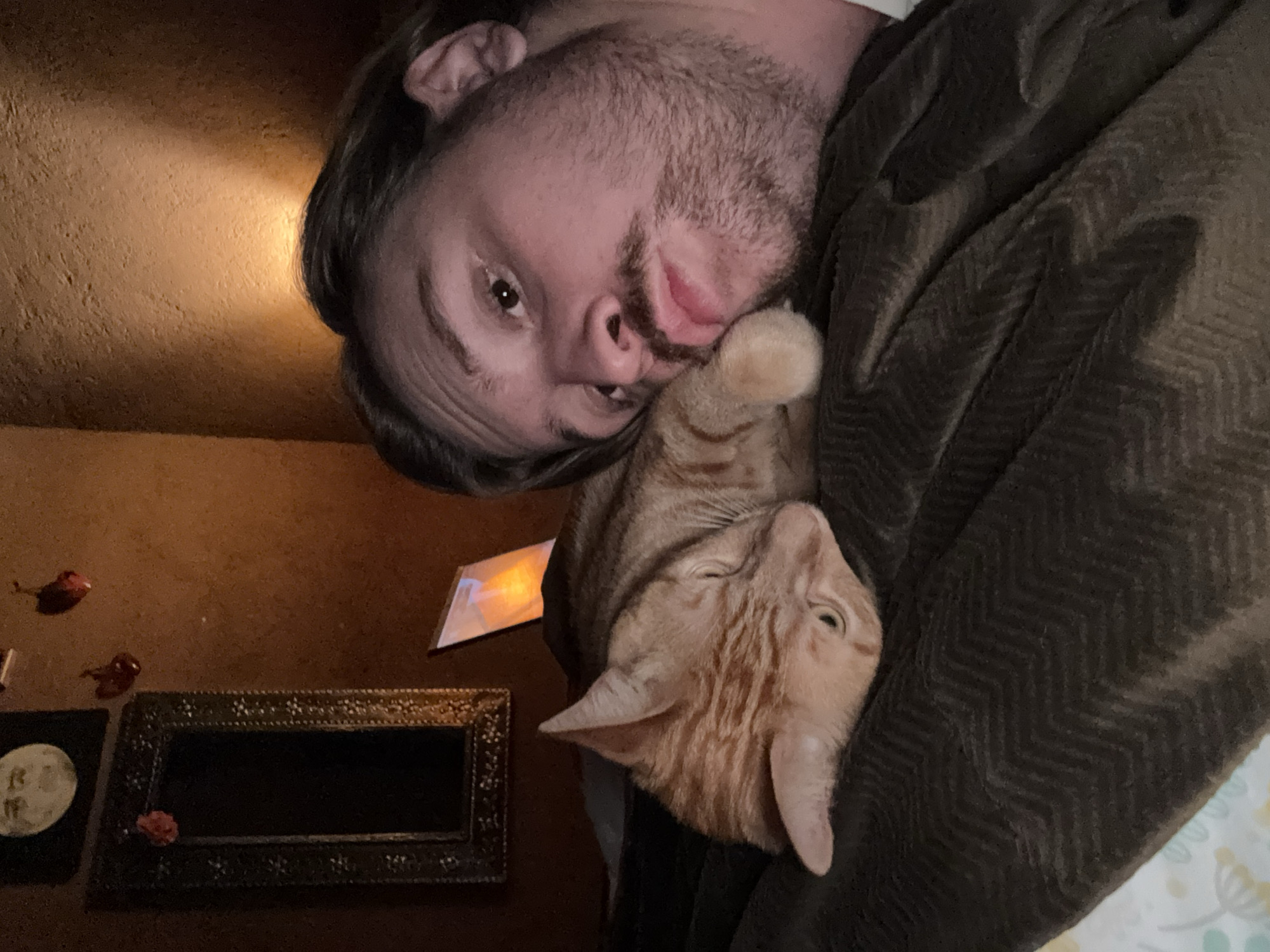 A man is seen from a low-angle perspective, holding an orange tabby cat against his chest. The man's face is close to the camera, and he has a slight beard and short brown hair. The cat is pressed against his cheek. They are partially covered by a brown blanket with a chevron pattern. In the background, a brown wall is visible, with a rectangular picture frame and other decorative items hanging on it. The location of the image is unknown and there is no verifiable evidence to indicate it was taken in the city of Cañada de Madero, Mexico.