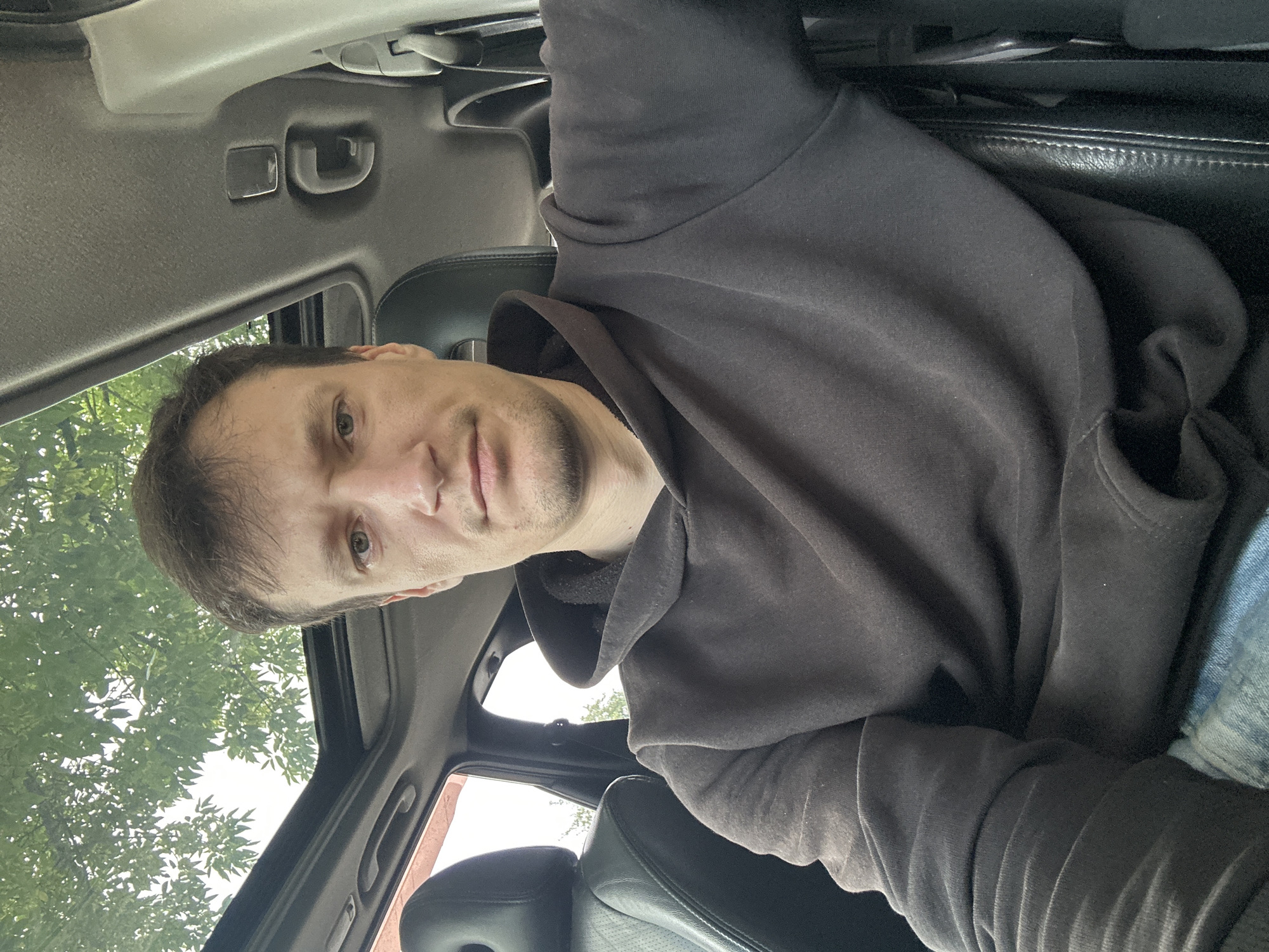 A man is seated inside a car, facing the camera. He has light brown hair and a fair complexion. He is wearing a dark brown hoodie and blue jeans. The car has a sunroof, through which green foliage from trees is visible. The interior of the car appears to be dark, with black leather seats and dark grey trim. The image was taken outdoors, likely in Makiv, Ukraine, given the context.