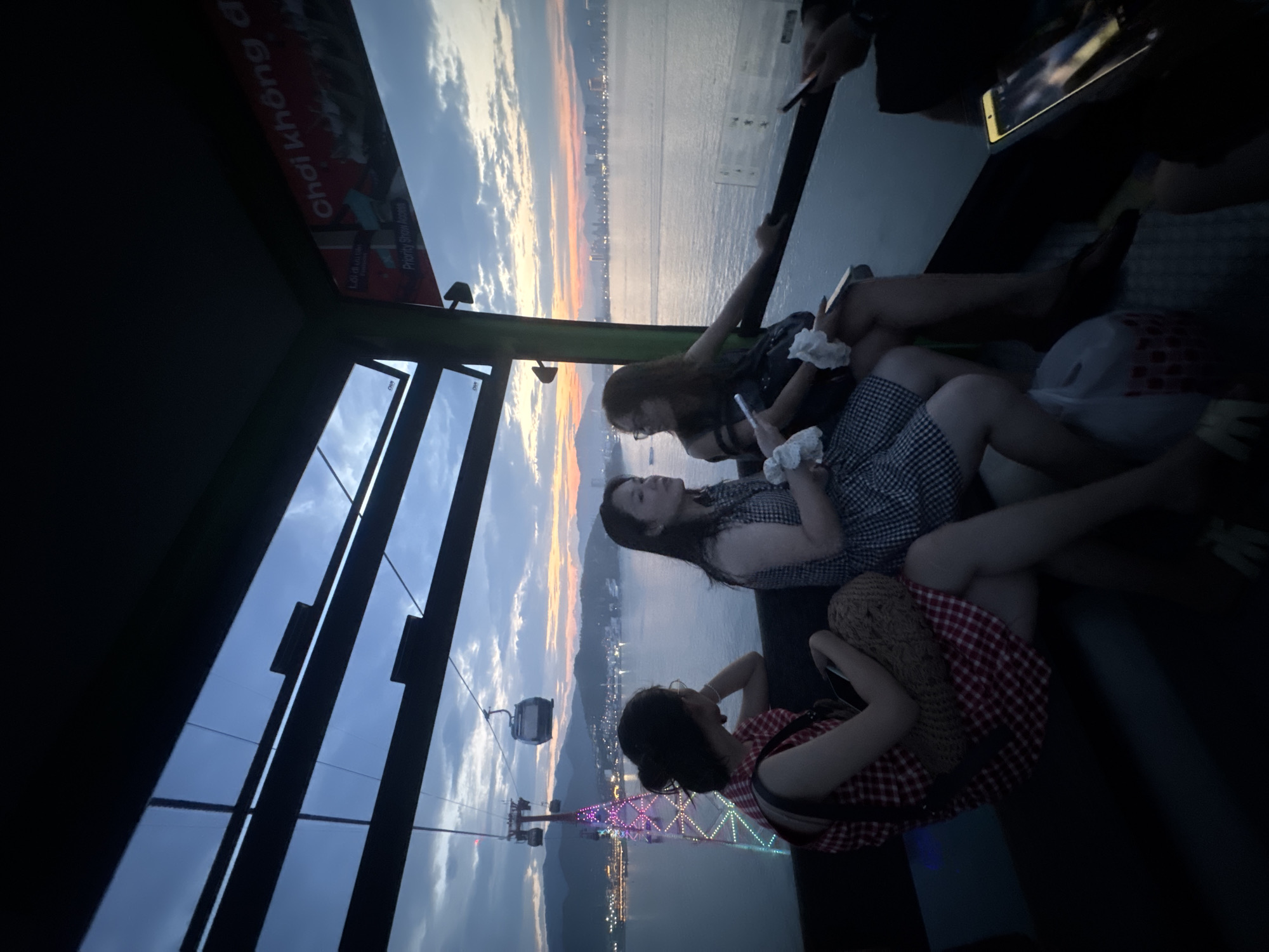 A group of three young women are seated inside a cable car traveling over the sea at dusk. The woman on the left wears a red and white checkered dress and carries a woven bag. The woman in the middle is in a black and white checkered dress, holding a white tissue. The woman on the right wears a dark colored dress and is also holding a white tissue. In the background, a brightly lit tower with colorful lights stands in the water, and the city lights of Nha Trang, Vietnam, are visible across the bay. The sky is a gradient of blues and oranges, indicating sunset. The cable car's interior is visible, with dark seats and a window offering a panoramic view.