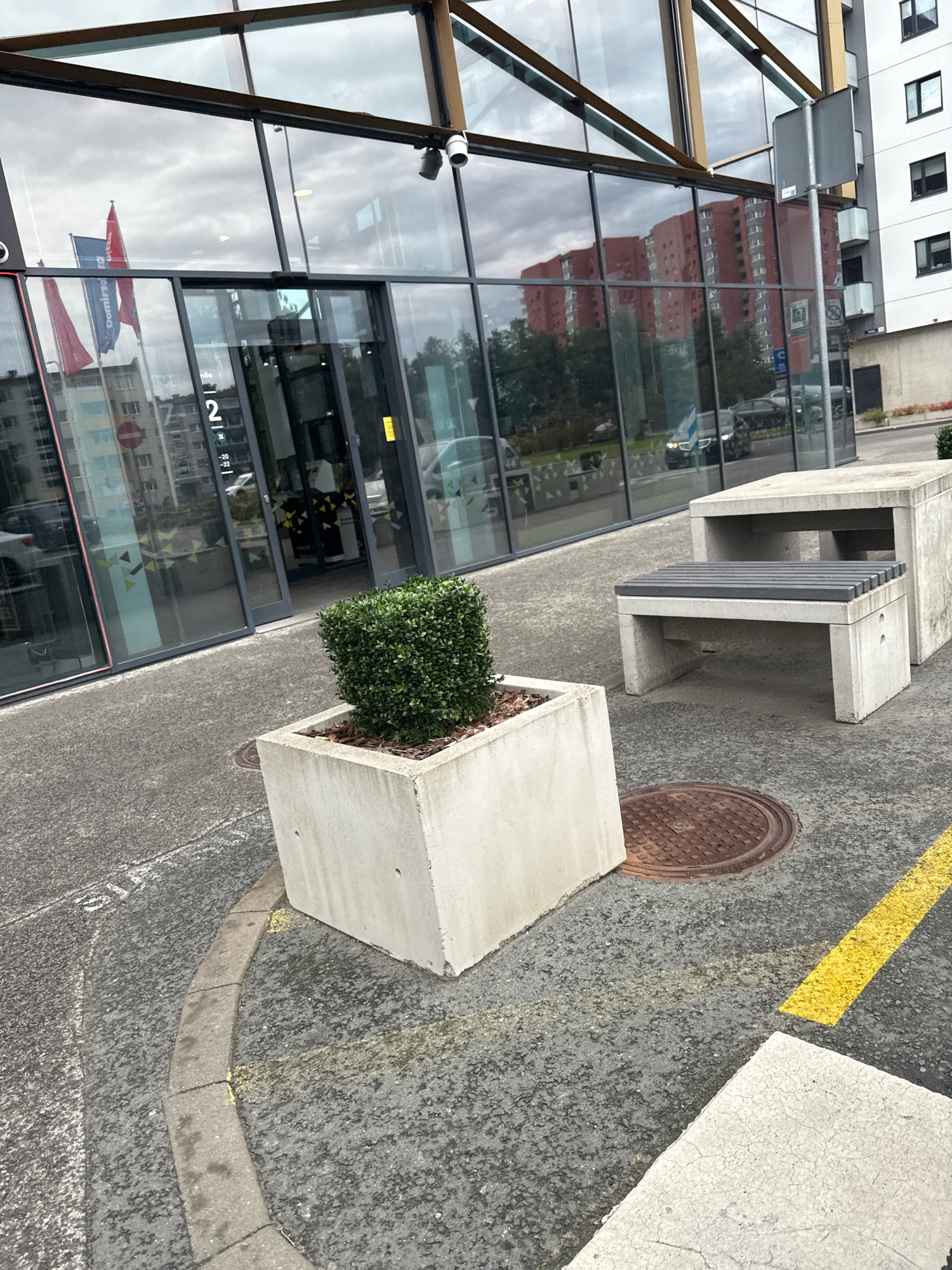 The image shows an outdoor scene in front of a modern building with a glass facade in Tallinn, Estonia. The building features large windows reflecting the cloudy sky and surrounding buildings. To the left, flags, one red and one blue with white lettering, are visible outside the building. In the foreground, a square concrete planter contains a neatly trimmed green bush. Further to the right, a concrete bench with a grey slatted seat is positioned next to a larger concrete block. A circular metal manhole cover is visible on the asphalt ground between the planter and the bench. Yellow lines, possibly road markings or parking indicators, are present on the asphalt. The environment appears to be an urban public space.