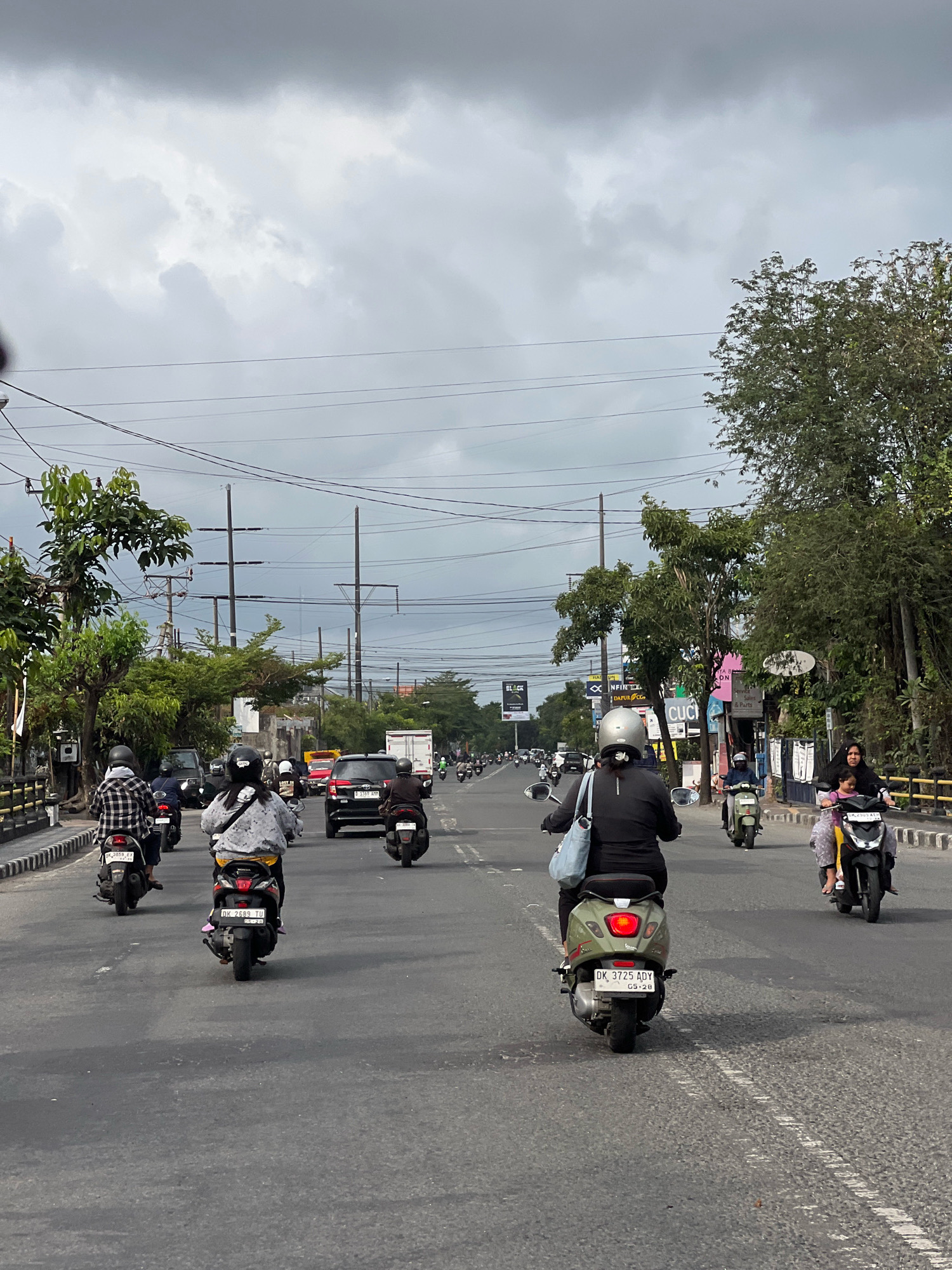 This image depicts a street scene in Denpasar, Indonesia, characterized by a cloudy sky and moderate traffic. The foreground shows several motorcycles and scooters on a paved road. One scooter in the center, ridden by a person wearing a black long-sleeved shirt and helmet, has a red taillight illuminated and a license plate reading "DK 3725 ADY" with "05-28" below it. To its right, a woman and a child on a yellow and black scooter are also visible. On the left, a person on a scooter with a checkered shirt and helmet is heading away from the camera, their license plate showing "DK 3859 EX". In the middle ground, more motorcycles, cars, and a white truck are present. The road is lined with trees and utility poles, with buildings and signs visible in the background. The overall atmosphere suggests a typical day in a busy urban environment.