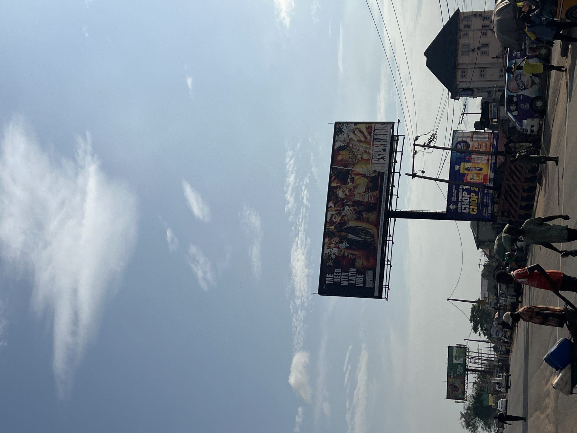This is a medium-shot, eye-level photograph taken on a sunny day in Onitsha, Nigeria. The scene captures a busy roadside with several billboards, vehicles, and people.
In the foreground, three men are walking along the side of the road. The man on the left is pushing a cart containing two large containers, one blue and one white. The middle man is wearing a red shirt and the man on the right is wearing a green shirt.
Further down the road, traffic is moving, with several cars and vans visible. Another billboard, advertising "INTUHA PRODUCTS CHOP 1 CHOP 2," is situated to the left of a multi-story building with a dark roof. Above this, a larger billboard for "DESPERADO" beer is prominently displayed against a blue sky with scattered clouds.
The environment appears to be a commercial area, with shops and vendors lining the street. Overhead power lines are visible, suggesting urban infrastructure. The overall impression is of a bustling, everyday scene in a Nigerian city.