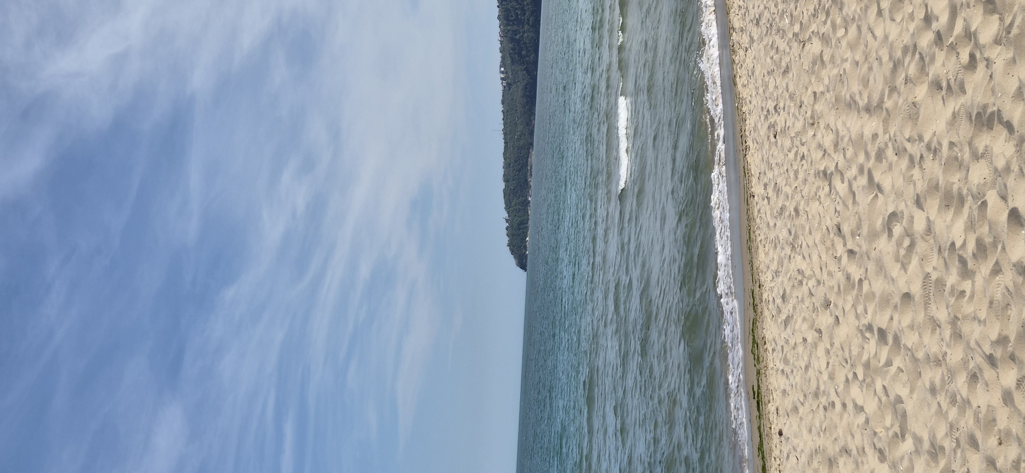 The image depicts a beach in Varna, Bulgaria. A sandy beach extends along the right edge of the frame, covered with footprints. Adjacent to the beach is the sea, with small waves visible near the shore. A green, tree-covered hill or cliff rises along the horizon line in the center of the image. Above, a blue sky is filled with wispy clouds. The composition is vertical.