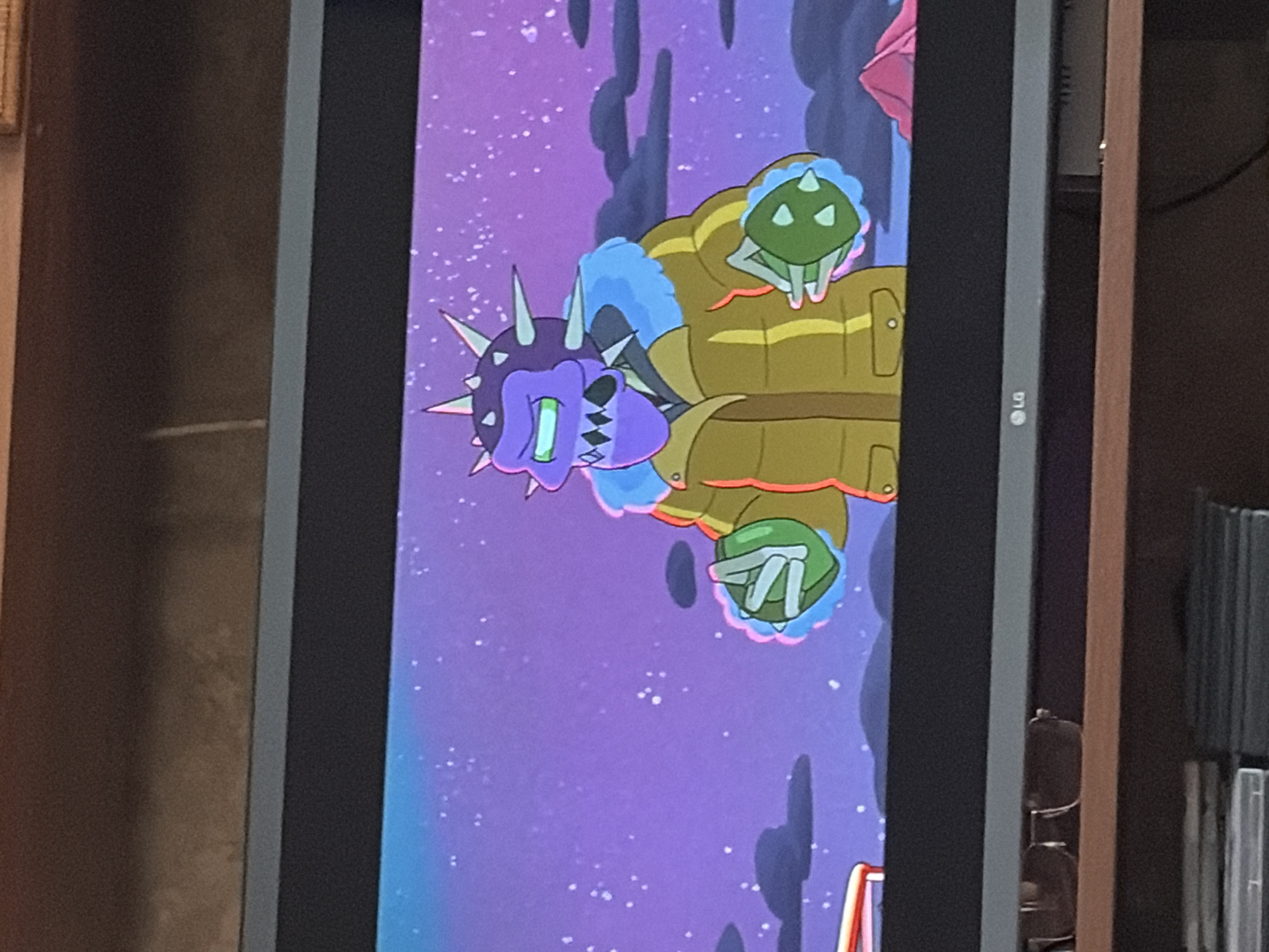 The image shows a cartoon character from the show Rick and Morty on a television screen. The character is purple with green eyes and spikes on its head. It is wearing a yellow jacket with blue fur trim and green spiked mittens. The background of the cartoon is a purple sky with white stars and dark clouds. The television screen is on a wooden shelf, and the top of the shelf has a pair of sunglasses on it. The bottom of the shelf has stacks of DVDs or Blu-rays. The scene takes place in an unknown city.
