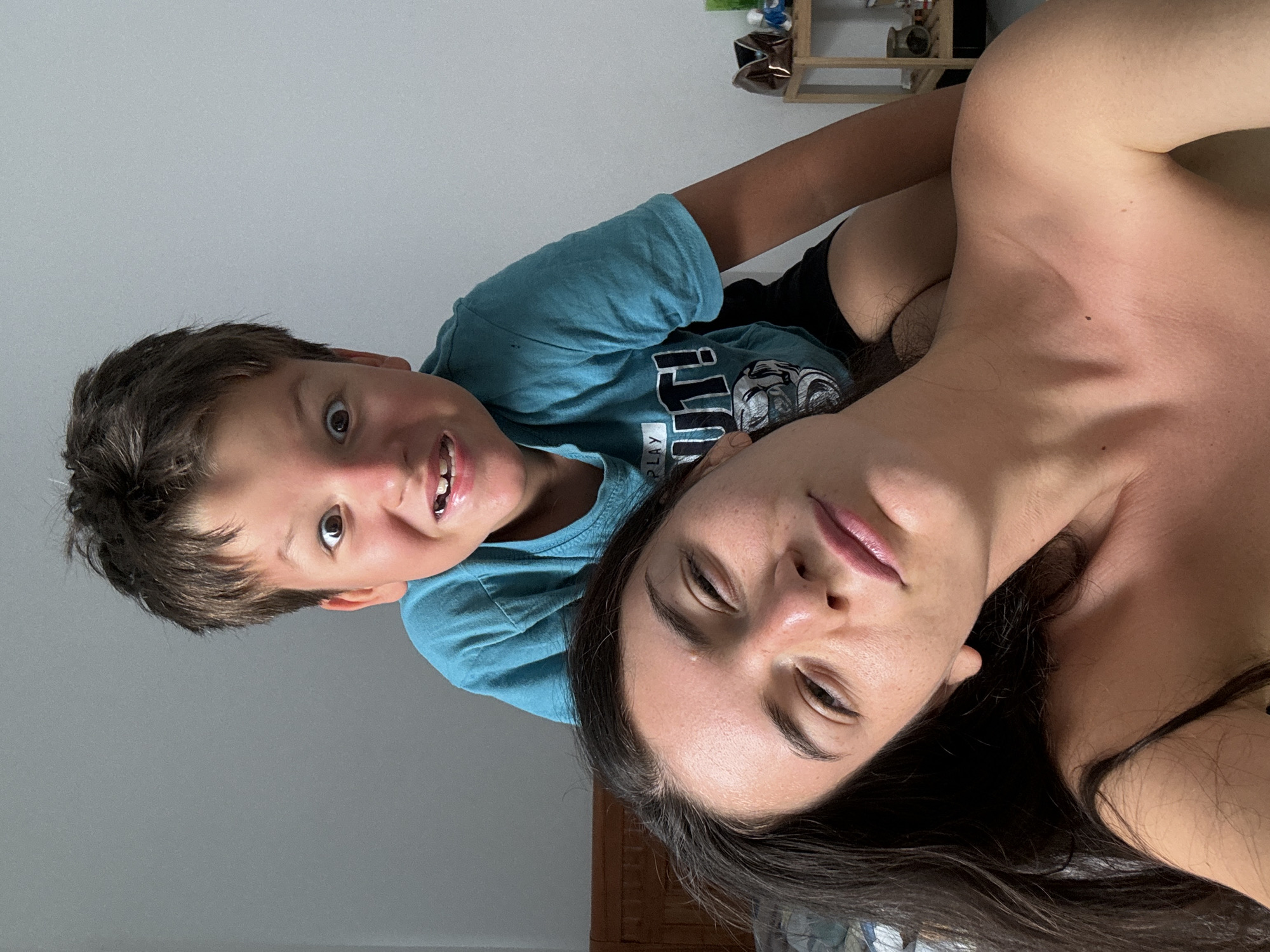 A close-up shot depicts a woman and a child, likely a mother and son, lying next to each other. The woman is facing the camera with her eyes partially closed, while the child is positioned above her, gazing directly at the camera with a broad smile. The woman has dark hair and appears to be partially unclothed. The child wears a teal t-shirt with a graphic on the front. The background includes a portion of a white wall and a wooden shelf or cabinet. There is no indication of location in the city of Mũi Né, Vietnam.