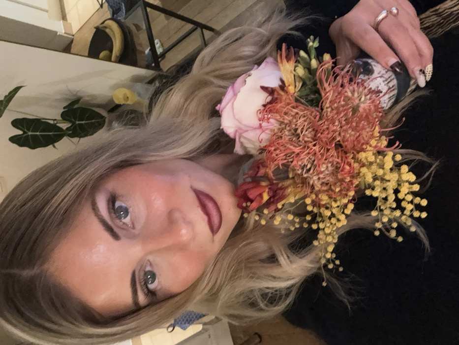 The media file presents a close-up portrait of a woman indoors, set in Paris, France. The woman, with long blonde hair and blue eyes, is looking directly at the viewer with a neutral to slightly content expression. She has distinct eyebrows and is wearing makeup, including eyeliner and dark red lipstick.
She is holding a vibrant bouquet of flowers near her chest and shoulder. The bouquet features a prominent light pink and white rose, several spiky reddish-orange flowers (possibly a variety of protea), numerous small yellow clustered flowers (resembling mimosa or baby's breath), and some reddish-orange closed bud-like flowers. Her left hand, adorned with a silver ring on her ring finger, is visible holding the flowers. Her fingernails are painted, some a dark color, and one with a white base and black polka dots. She appears to be wearing a dark-colored top.
The background is dimly lit, suggesting an indoor setting, likely in the evening or at night. Elements in the background include a dark green houseplant with broad leaves and a blurred glimpse of a domestic interior, possibly a shelf or table with some yellow objects (perhaps fruit like bananas) and other household items. No discernible text is visible in the image.