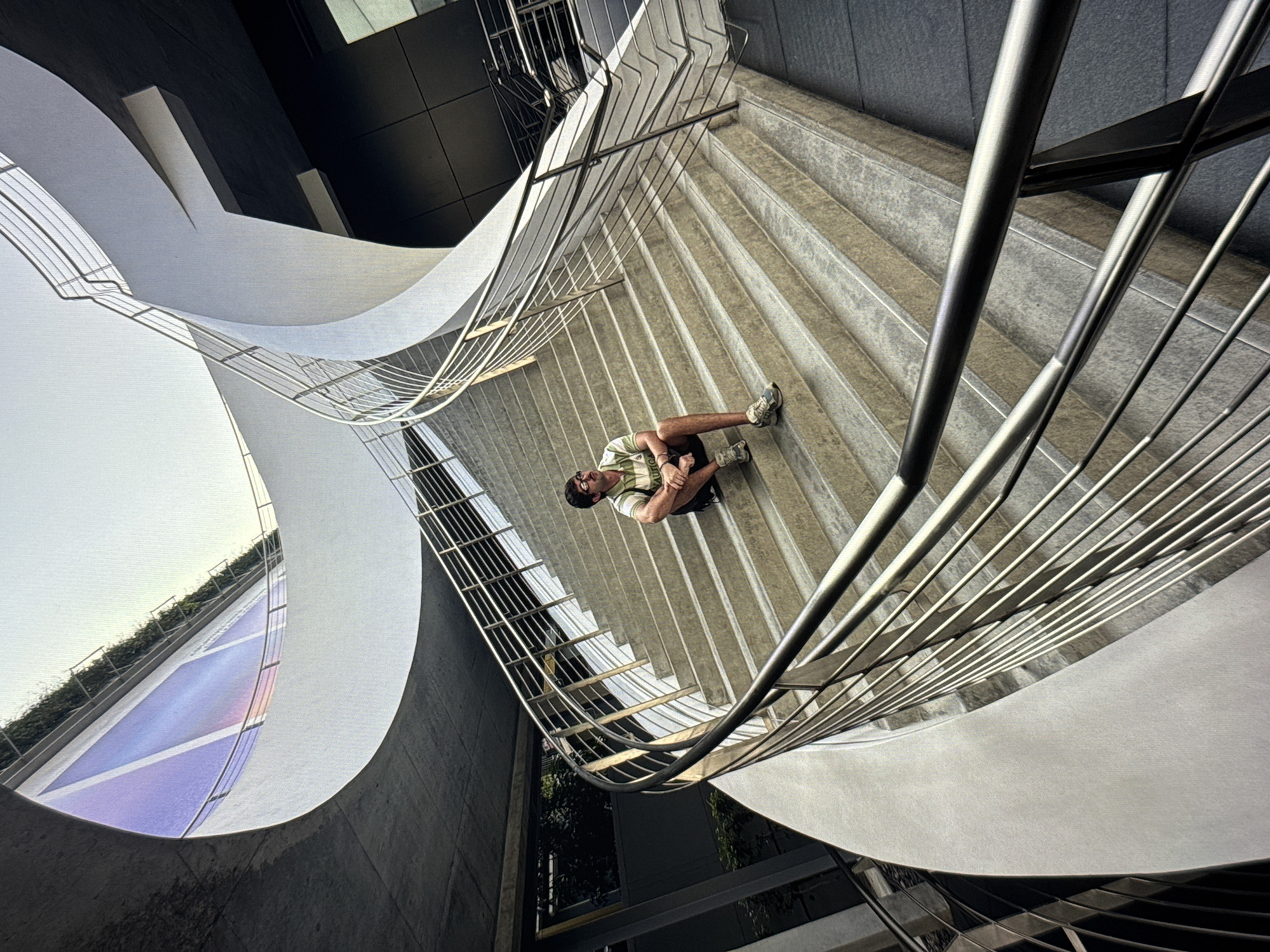 A person sits on concrete steps of a modern, curved outdoor staircase. The person is a male wearing a green and white striped t-shirt, dark shorts, and athletic shoes, with glasses. He is looking upwards and to the left. The staircase has metal railings. In the background, there is a large digital billboard with abstract colorful graphics and some greenery on an elevated walkway. The architecture appears contemporary, with smooth white concrete elements and dark building facades. The lighting suggests daylight. The location is not identifiable as Moscow, Russia.