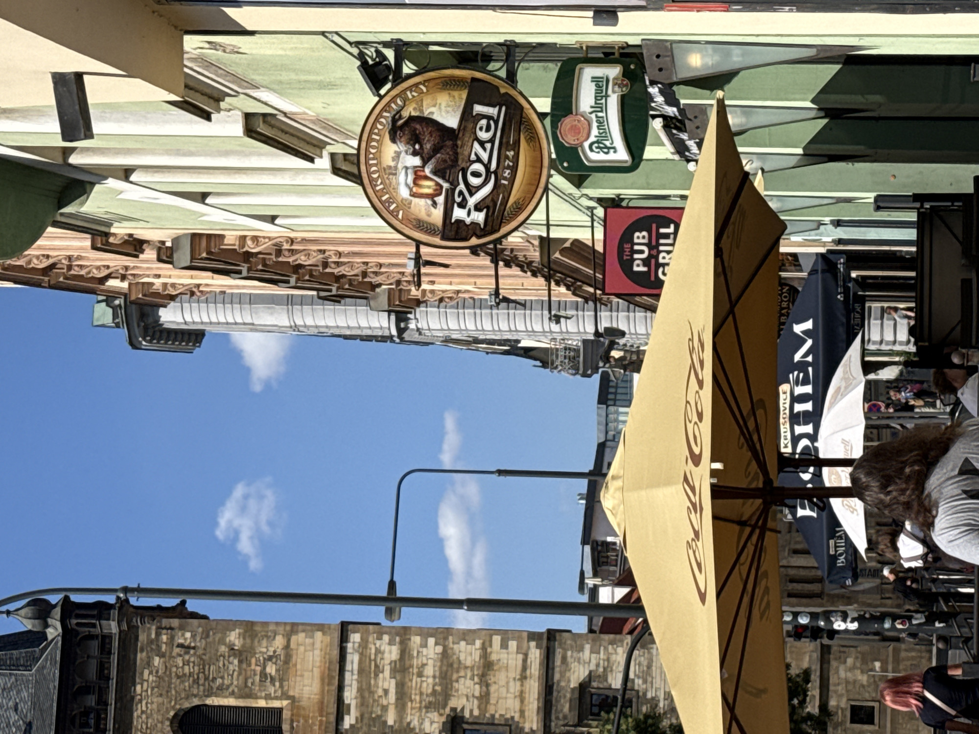 The image captures a street scene in Prague, Czechia, on a clear, sunny day. In the foreground, a large yellow umbrella, branded with "Coca-Cola," partially obscures the view of several awnings and umbrellas further back. One awning displays the word "KRUSOVICE" and the name "BOHÉM," while another features "THE PUB & GRILL." A light-colored umbrella with "Well" printed on it is also visible.
Dominating the mid-ground is a round sign advertising "Kozel" beer, featuring an illustration of a goat holding a beer mug. Below this, a green rectangular sign reads "Pilsner Urquell." Behind these signs, a light green building with cream-colored pillars and decorative trim runs along the street. To the left, a stone building with an arched window and a tall, slender spire-like structure is visible, likely part of a historic church or tower. Two streetlights with angular arms are also present.
In the background, the facades of various buildings with distinct architectural styles line the street. Several individuals are present, though their features are not clearly discernible. One person in the foreground on the left has pink hair and wears a black top and a crossbody bag. Further back, several people are seen near the awnings and umbrellas, some appearing to be patrons or staff. The overall impression is that of a vibrant outdoor seating area outside establishments in a historic European city.
