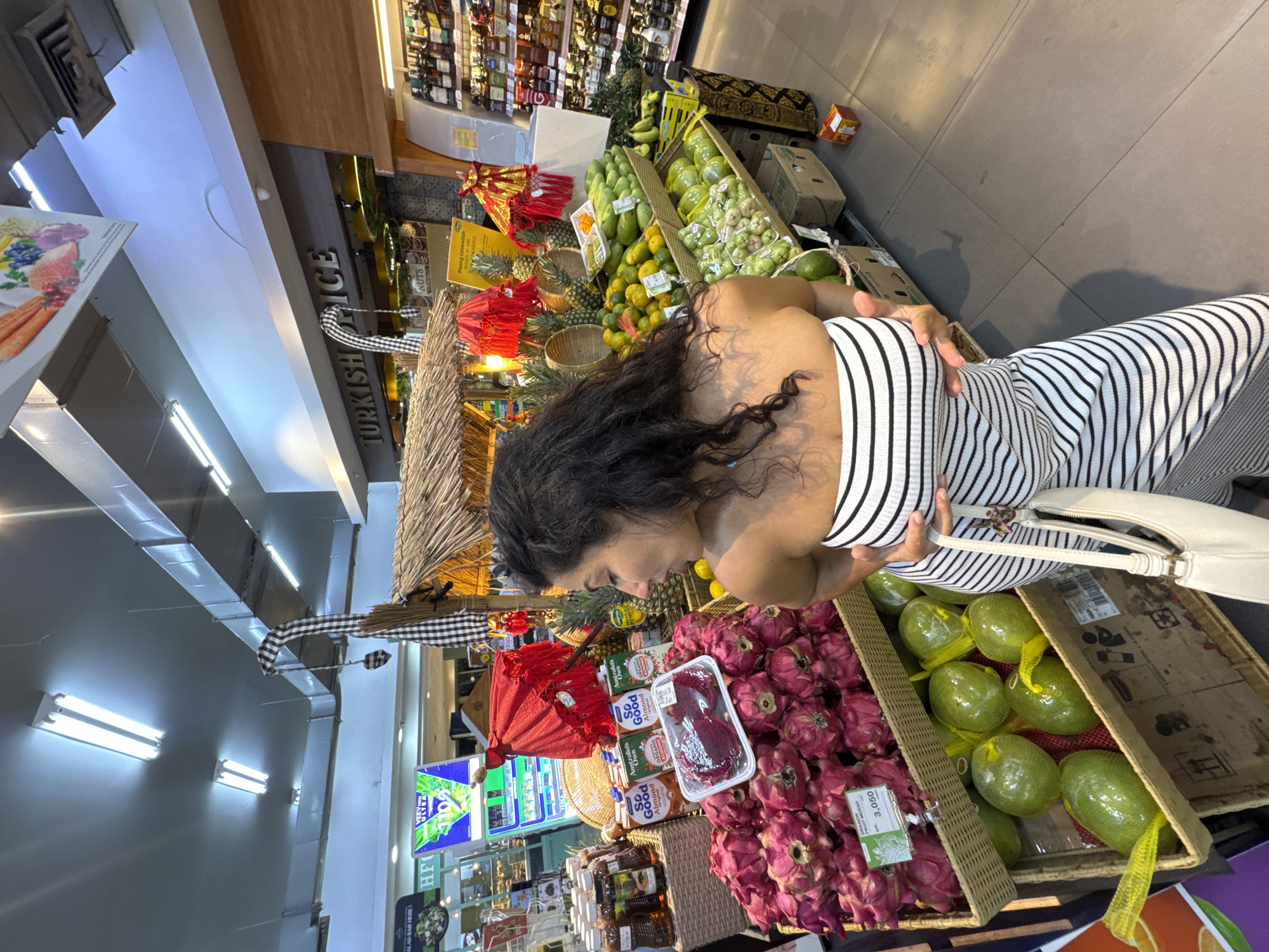 The image captures a woman browsing a fruit and produce section inside what appears to be a supermarket or grocery store in North Kuta, Indonesia. The woman, with dark, curly hair, is seen from a slightly elevated, over-the-shoulder perspective, wearing a black and white striped top or dress. She is intently looking down at a vibrant display of tropical fruits.
In the foreground, large quantities of pinkish-red dragon fruit ("DRAGON FRUIT MERAH") are displayed in a woven basket, with a price tag showing "3,050" (likely Indonesian Rupiah, perhaps per 100g or per fruit if 30,500 IDR). Next to it, another basket holds several green, oval-shaped fruits, each individually netted in yellow mesh, possibly mangoes or similar. Further displays behind the woman feature numerous green mangoes, yellow-orange citrus fruits (possibly oranges or tangerines), pineapples, and smaller clusters of light green, bell-shaped fruits (potentially rose apples or water apples). Some of the fruit displays are arranged under thatched roofs, giving a market stall aesthetic within the store.
Beyond the fruit section, various packaged goods are visible on shelves, including "So Good Australia's Own Organic Almond" milk cartons and jars of what look like preserves or sauces. A sign with "NUTS" is visible. On a yellow sign, the word "Pisang" (Indonesian for banana) is partially visible. Further back, a sign reads "TURKISH SPICE" indicating another specialty section. The ceiling is lined with fluorescent lights, illuminating the store brightly, suggesting daytime. Another sign in the background reads "SALAD BAR HERE!". The overall scene depicts a busy and well-stocked grocery environment, emphasizing fresh produce and specialty items.