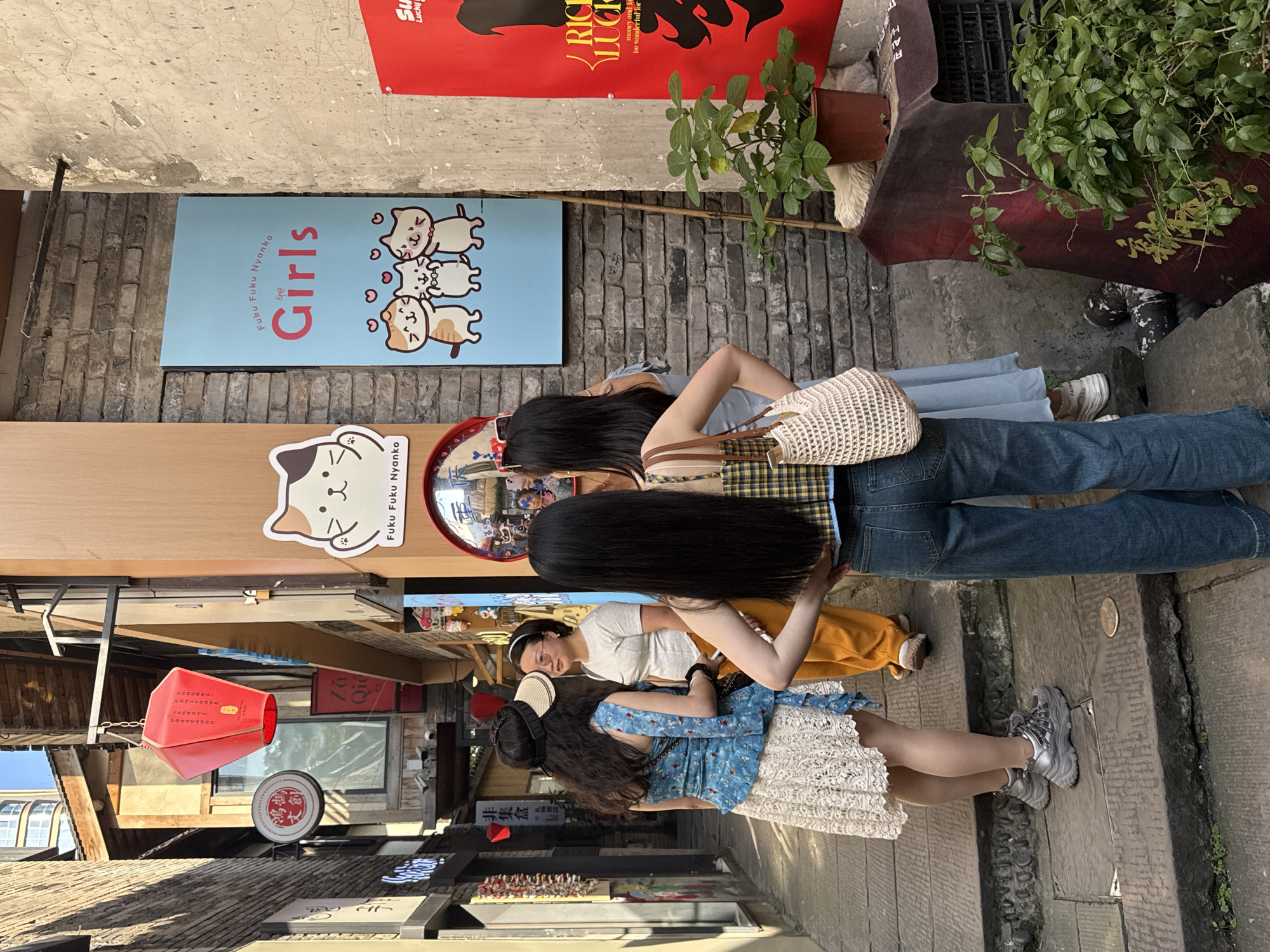 Three young women are casually walking or standing in a narrow, sunlit alleyway, likely a bustling street market or shopping area in Yuzhong District, China. The woman in the foreground has long black hair, a plaid top, blue jeans, and carries a woven bag, facing away from the camera. Beside her, another woman is partially visible wearing a grey t-shirt and yellow pants. In front of them, a third woman with long dark hair is seen from behind, dressed in a blue patterned top, a white lace skirt, and chunky grey sneakers.
The alley is paved with stone or brick, and lined with various shops and buildings with traditional architectural elements. Bright sunlight illuminates the scene, suggesting daytime.
Visible signs and objects include a light blue sign on a wall featuring three cartoon cats and the word "Girls" along with "Fuku Fuku Nyanko." Another white sticker with "Fuku Fuku Nyanko" is on a wooden beam. A red sign partially visible at the top advertises "RICH LUCK" with the word "Lucky" and phrases like "money and lo" and "too wonderful for." Other shop signs include "Za Qid" and Chinese characters like "非集盒" (fēi jí hé) and "文創" (wén chuàng, cultural and creative). Two potted plants are placed along the wall. A red, megaphone-like object hangs from an awning further down the street.