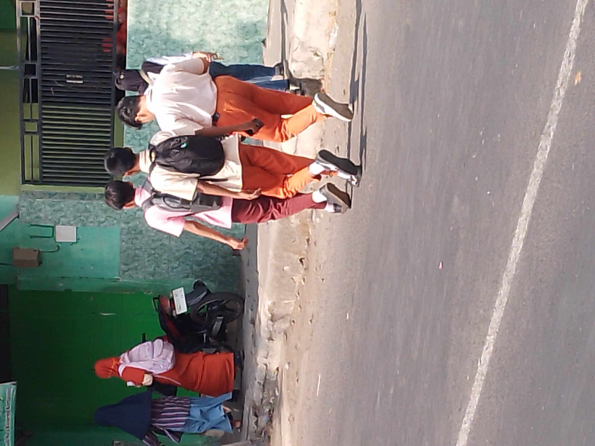 The image depicts a street scene in Kertosono, Indonesia. In the foreground, a paved road with a white dividing line is visible. On the right side of the road, a group of individuals are walking away from the camera. Three young males, seemingly students, are walking in single file. The one on the left is wearing a pink shirt and maroon pants, with a backpack. The middle student is in a tan shirt and orange pants, also with a backpack. The student on the right is wearing a light-colored shirt and orange pants, carrying a mobile phone. Behind them, a person in a blue shirt and dark pants is visible. Further in the background, on the left side of the image, two females are walking. One is wearing a dark blue hijab and a striped shirt with light blue pants, and the other is in an orange hijab and an orange dress, carrying a light pink backpack. A black motorcycle is parked near them, leaning on its kickstand. The background includes green walls, a metal fence, and a building. The overall scene appears to be a typical daytime street in a residential or school area of Kertosono.