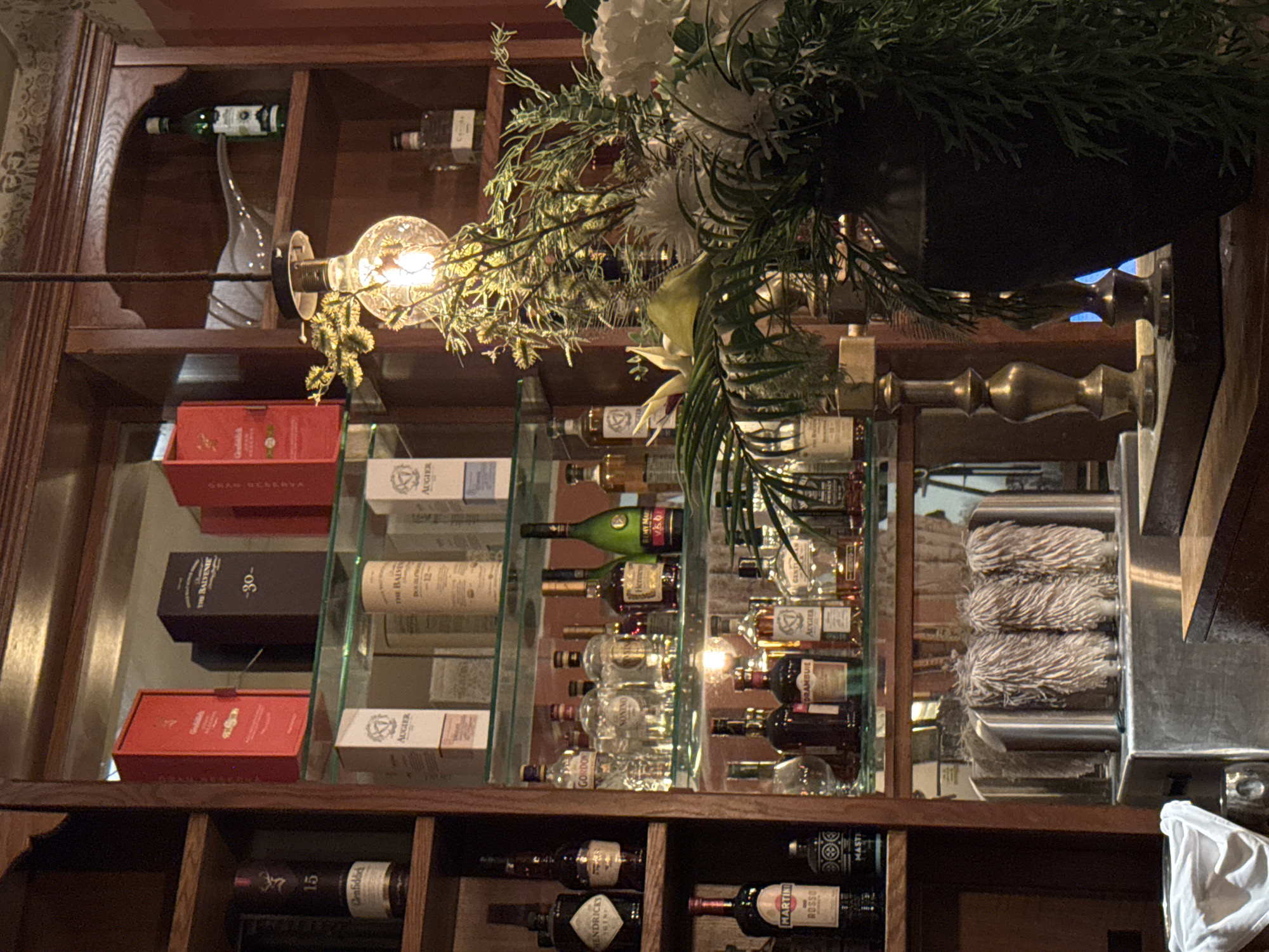 Here is a description of the image:
The image shows a well-stocked bar shelf, likely inside a restaurant or bar establishment. The shelves are made of dark wood and contain a variety of alcoholic beverages including whiskies, vodkas, and other liquors. Various brands are visible like Glenfiddich, Hendrick's Gin, Martini Rosso and Remy Martin. There is a glass shelf in the middle of the wooden shelves. There are decorative elements like flowers and greenery arranged around the shelf. A light fixture is attached to the shelf, illuminating the bottles. The reflection of the bottles is visible in the glass.
Based on the brands of alcohol and the overall presentation, the image most likely depicts a sophisticated bar setup. The location in Tel-Aviv, Israel, is not evident from the image.