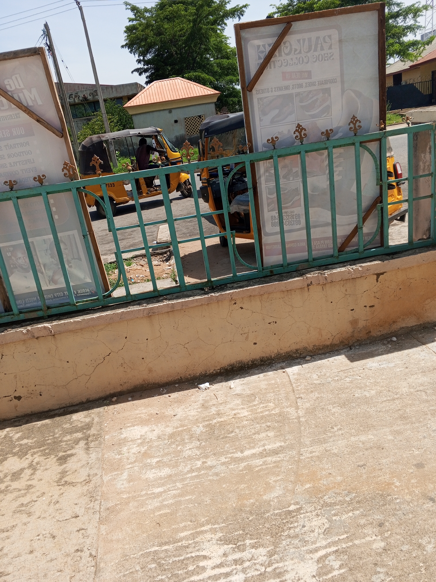 The image depicts a street scene in Jalingo, Nigeria, featuring a yellow auto-rickshaw with the number 7533 prominently displayed on its side. A person is seated in the driver's position of the auto-rickshaw. Two large advertisements are visible, one on the left and another on the right, leaning against a turquoise metal fence. The advertisement on the right is for "PAULLOGAN SHOE COLLECTION." The scene is set against a backdrop of trees and buildings under a bright, clear sky. The foreground consists of a concrete pavement and a yellow, cracked wall.